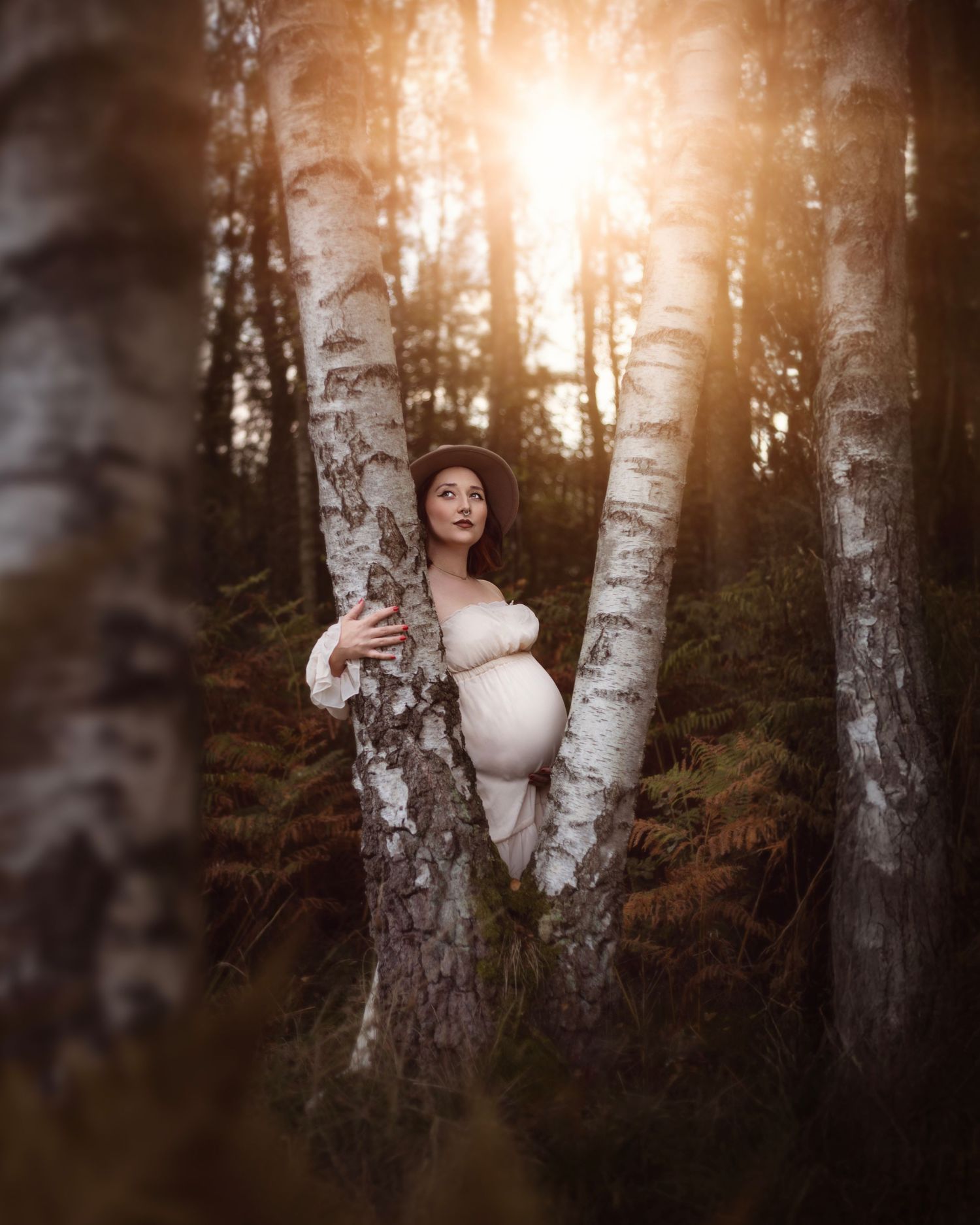 Pregnant woman in cream dress embracing birch trees in an enchanted forest at golden sunset in the West Midlands