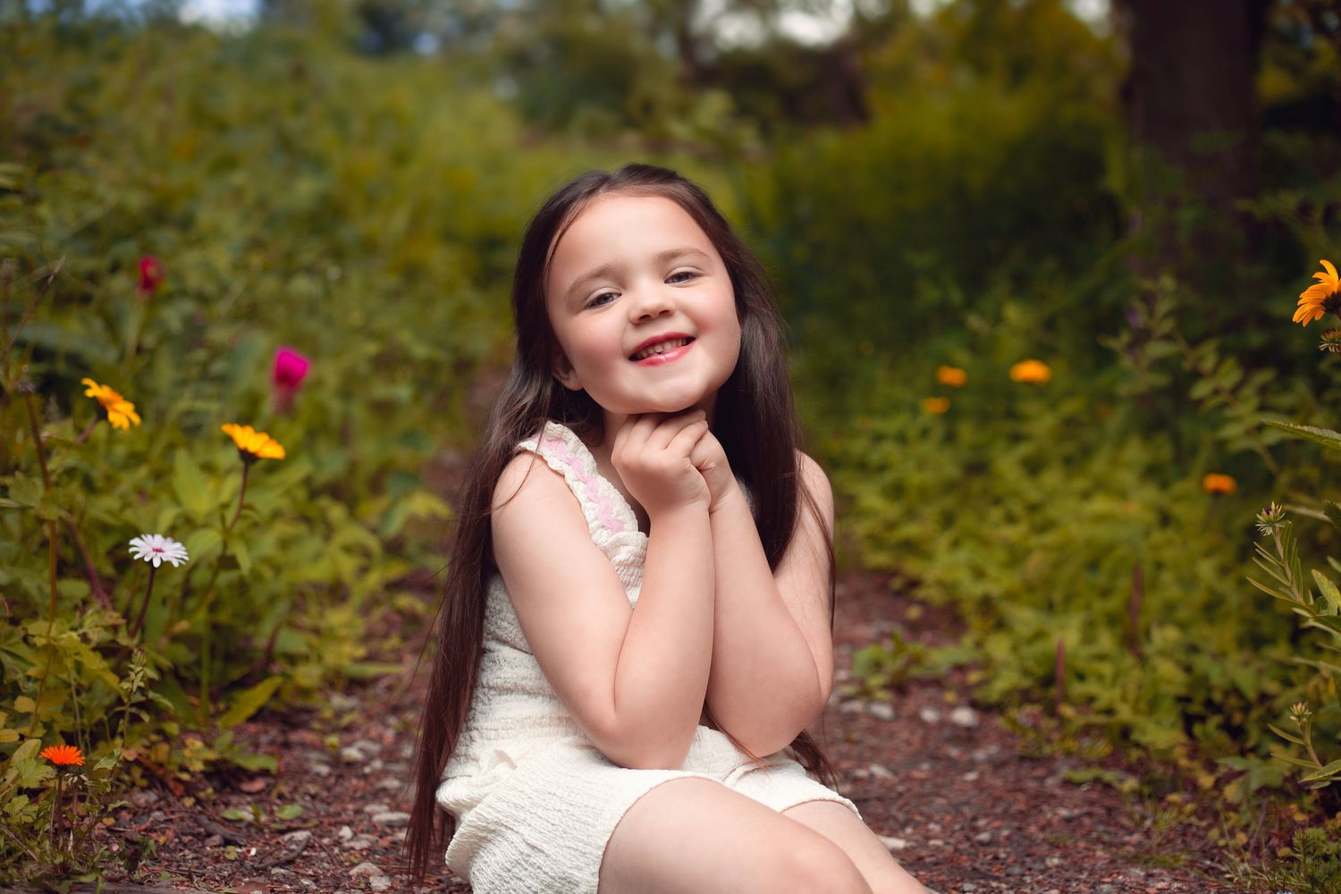 Smiling young girl with long dark hair sits among colorful wildflowers in a sunny garden setting, in the West Midlands