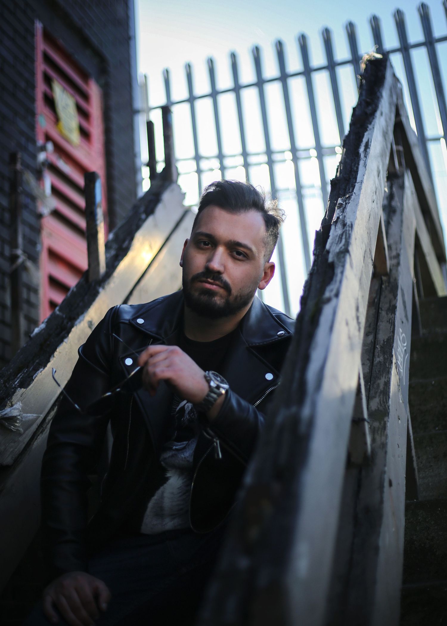 Bearded man in black leather jacket posing among weathered wooden planks in urban alleyway setting in the West Midlands