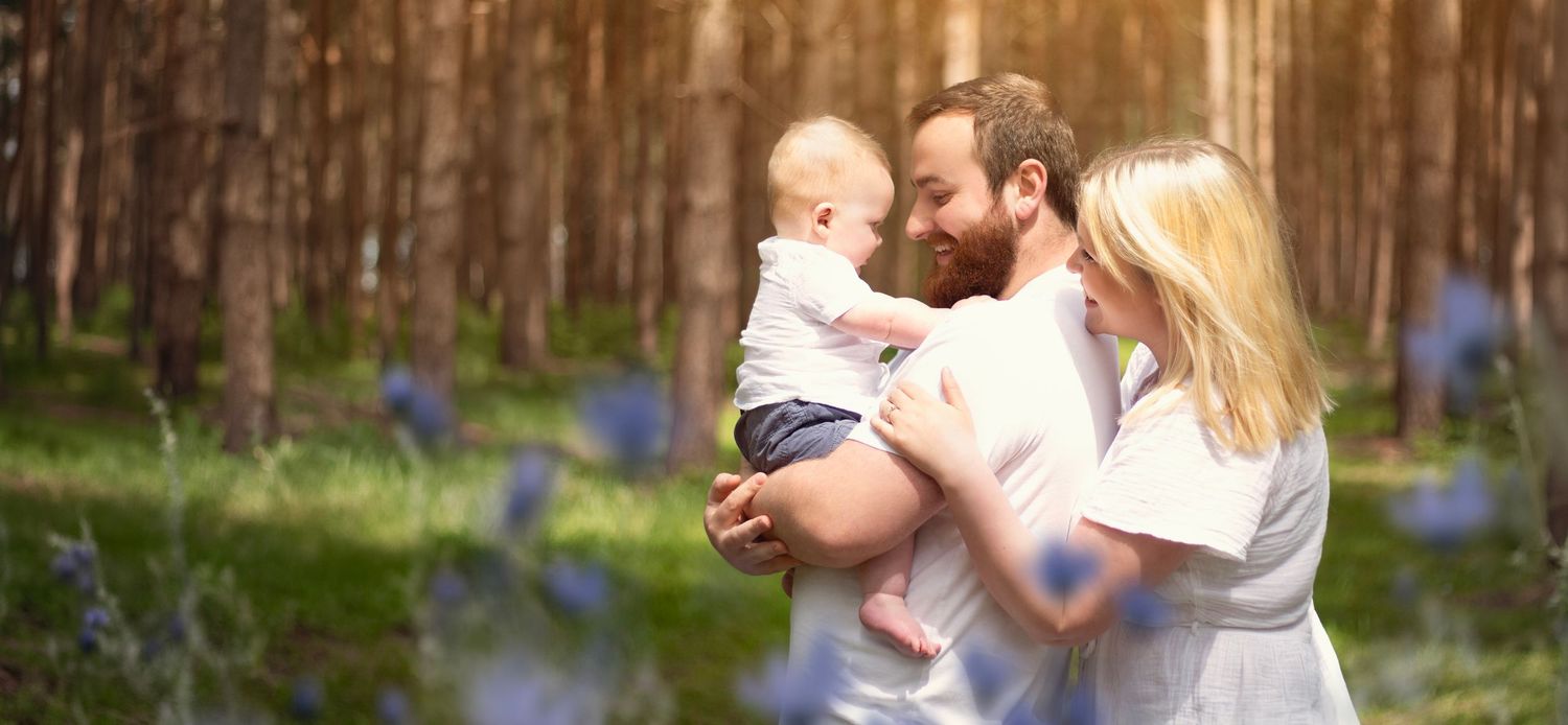 A young family with a baby shares a tender moment in a sunlit forest with blooming wildflowers  in Birmingham