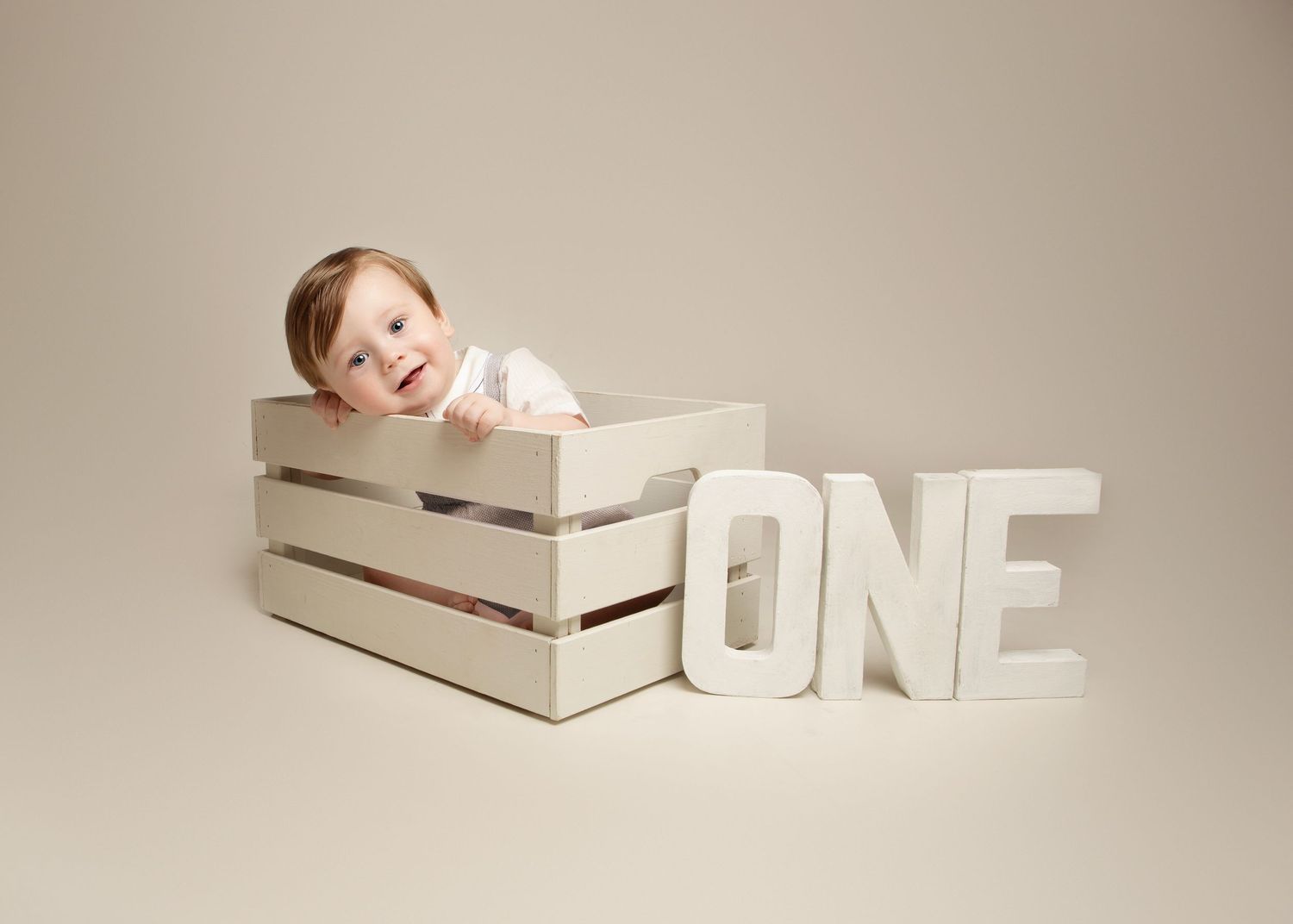 Smiling baby leaning over edge of white wooden crate next to ONE letter sign for first birthday photo in the West Midlands