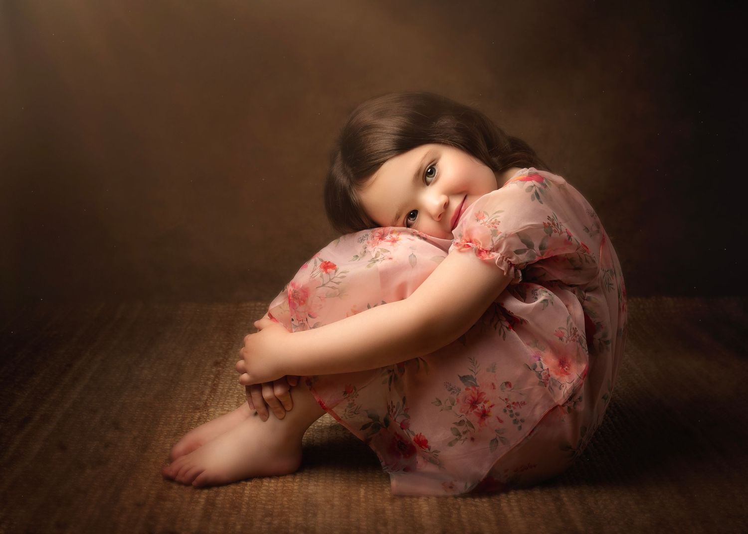 Adorable little girl in floral pink dress sits hugging knees on wooden floor in studio  in Birmingham