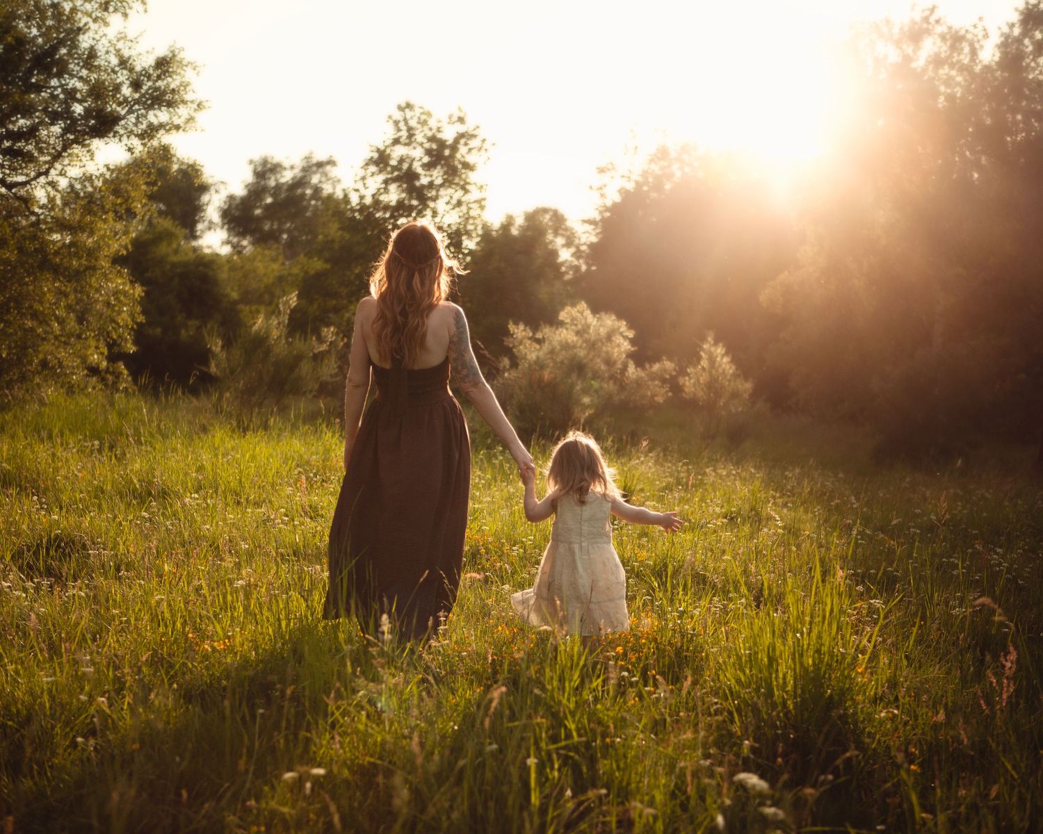 A mother and young daughter hold hands walking through a sunlit green meadow at golden hour.