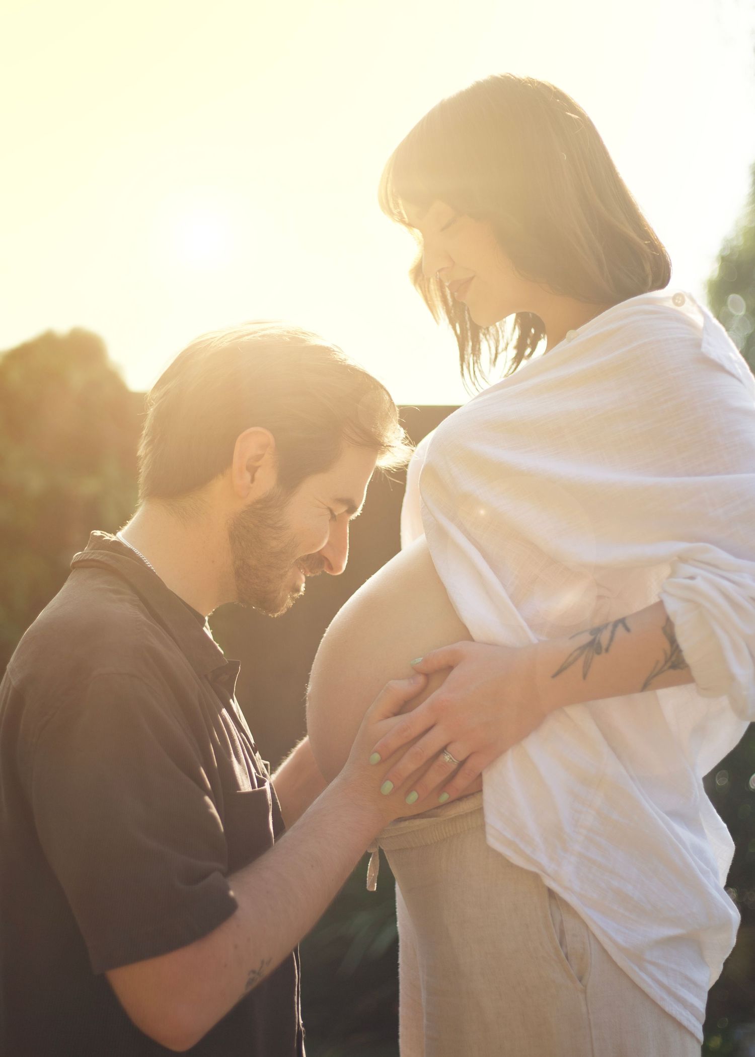 Man kneeling to touch pregnant partners baby bump in warm golden sunset light outdoors in the West Midlands