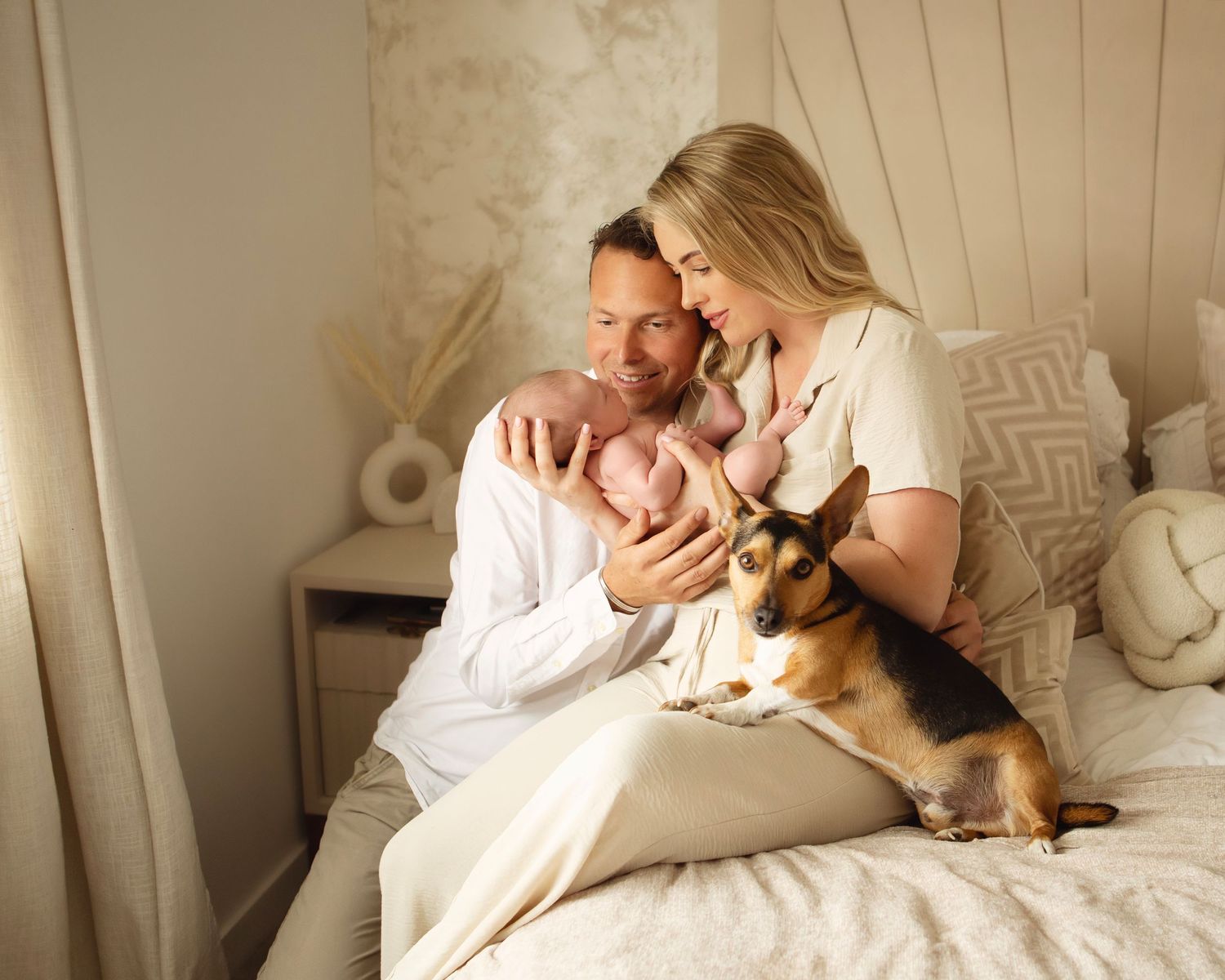 New parents sitting on bed with newborn baby and German Shepherd dog in cozy bedroom setting.