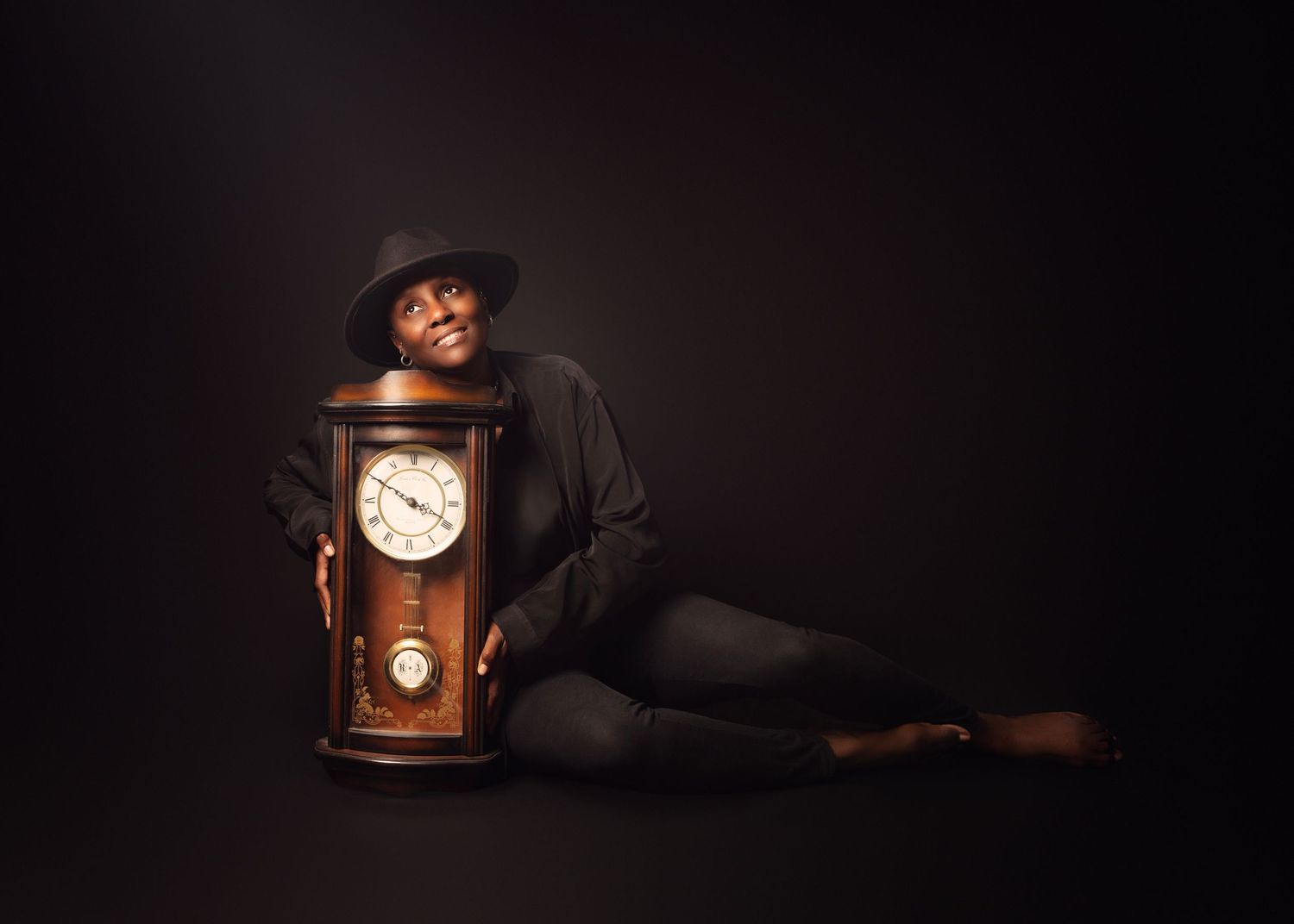 Person in black hat leans against a vintage wooden grandfather clock in a dramatic dark studio portrait, in Wolverhampton