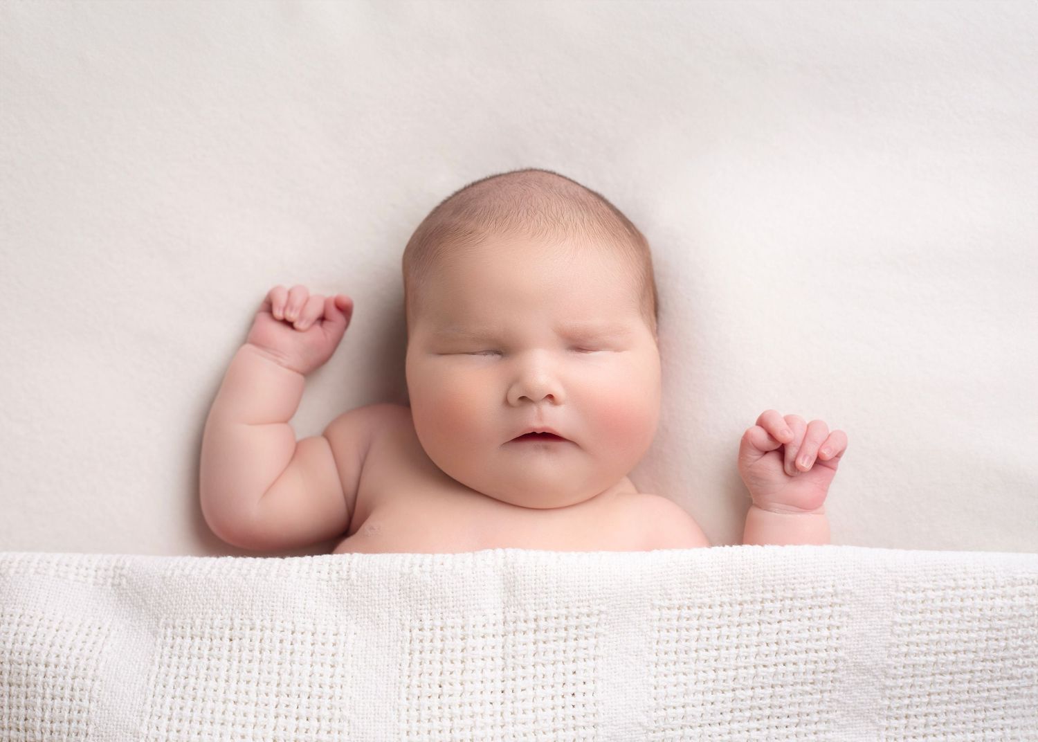 Peaceful newborn baby lying on a white blanket with tiny fists raised in the air in the West Midlands