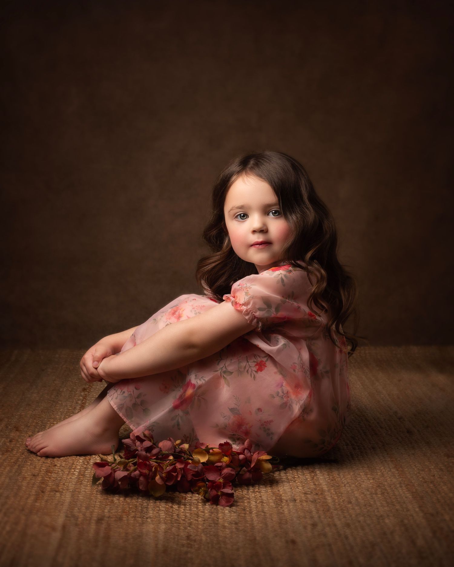 Beautiful little girl in pink floral dress sitting on floor with autumn leaves in studio portrait in the West Midlands