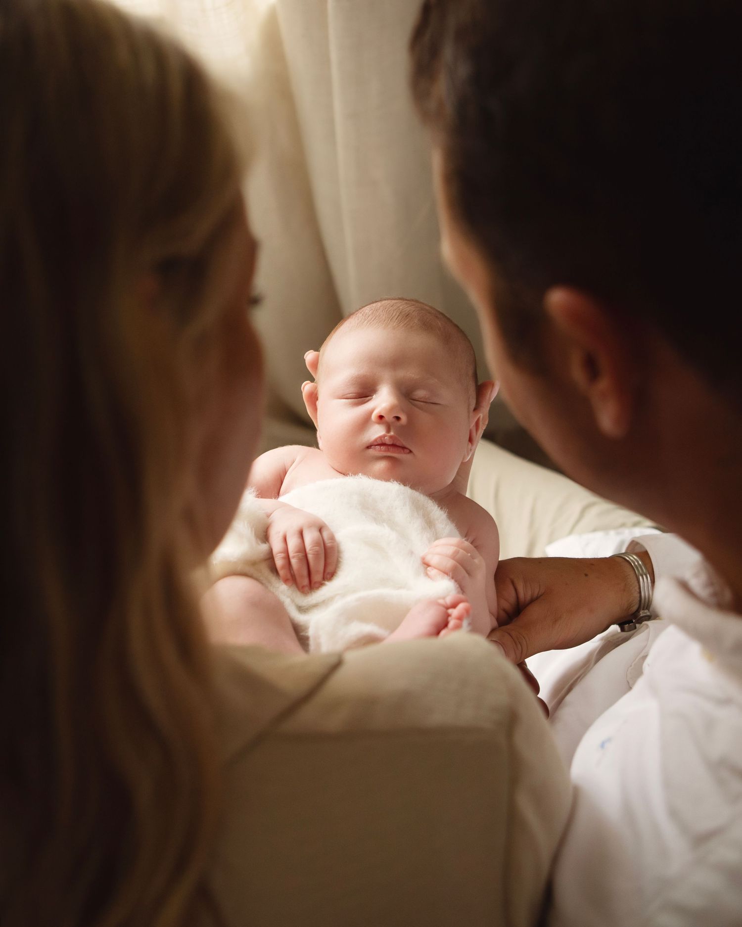Two parents tenderly gazing at their sleeping newborn baby wrapped in white cloth.