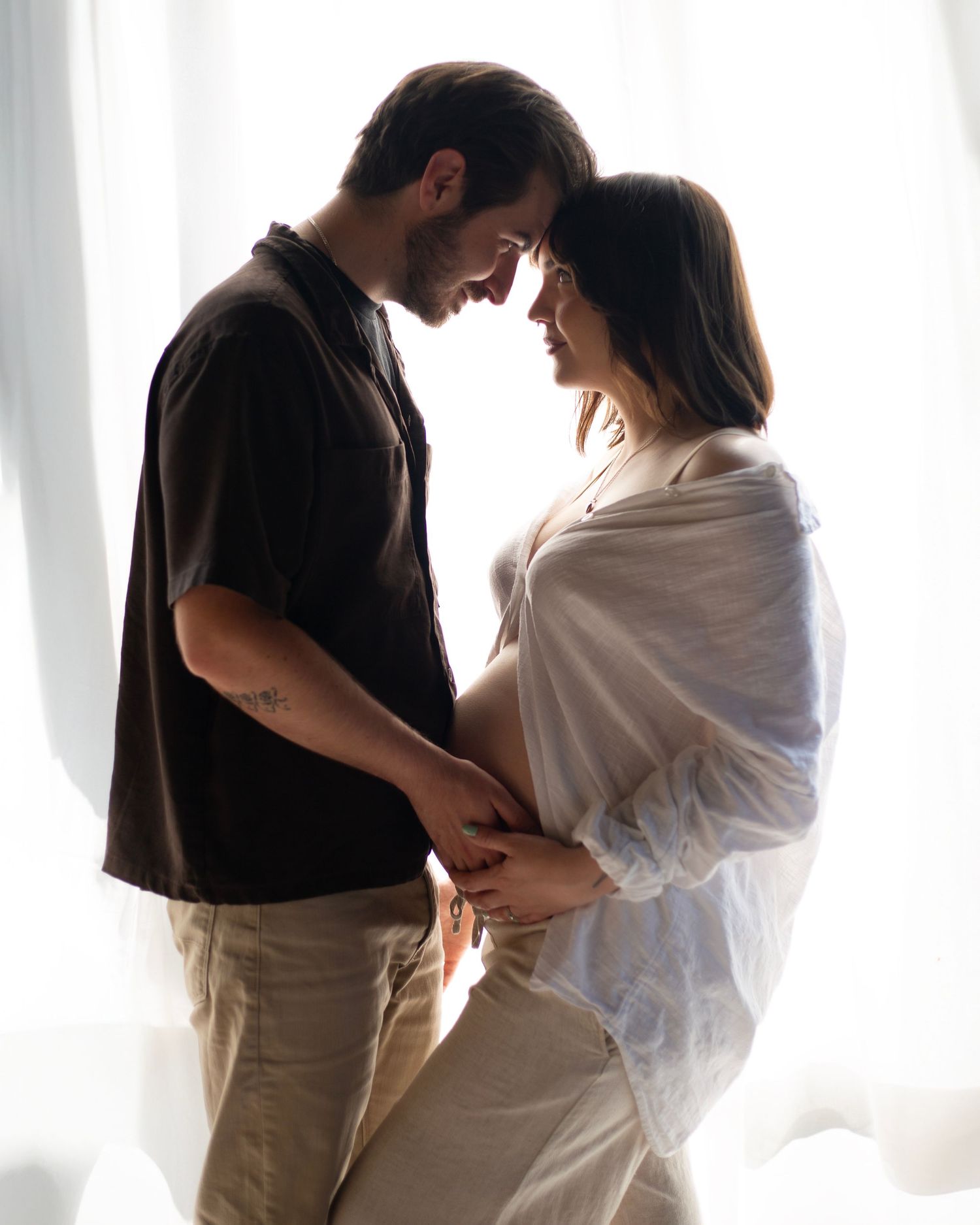 Couple sharing intimate moment hands on pregnant belly backlit by soft white curtains indoors in the West Midlands