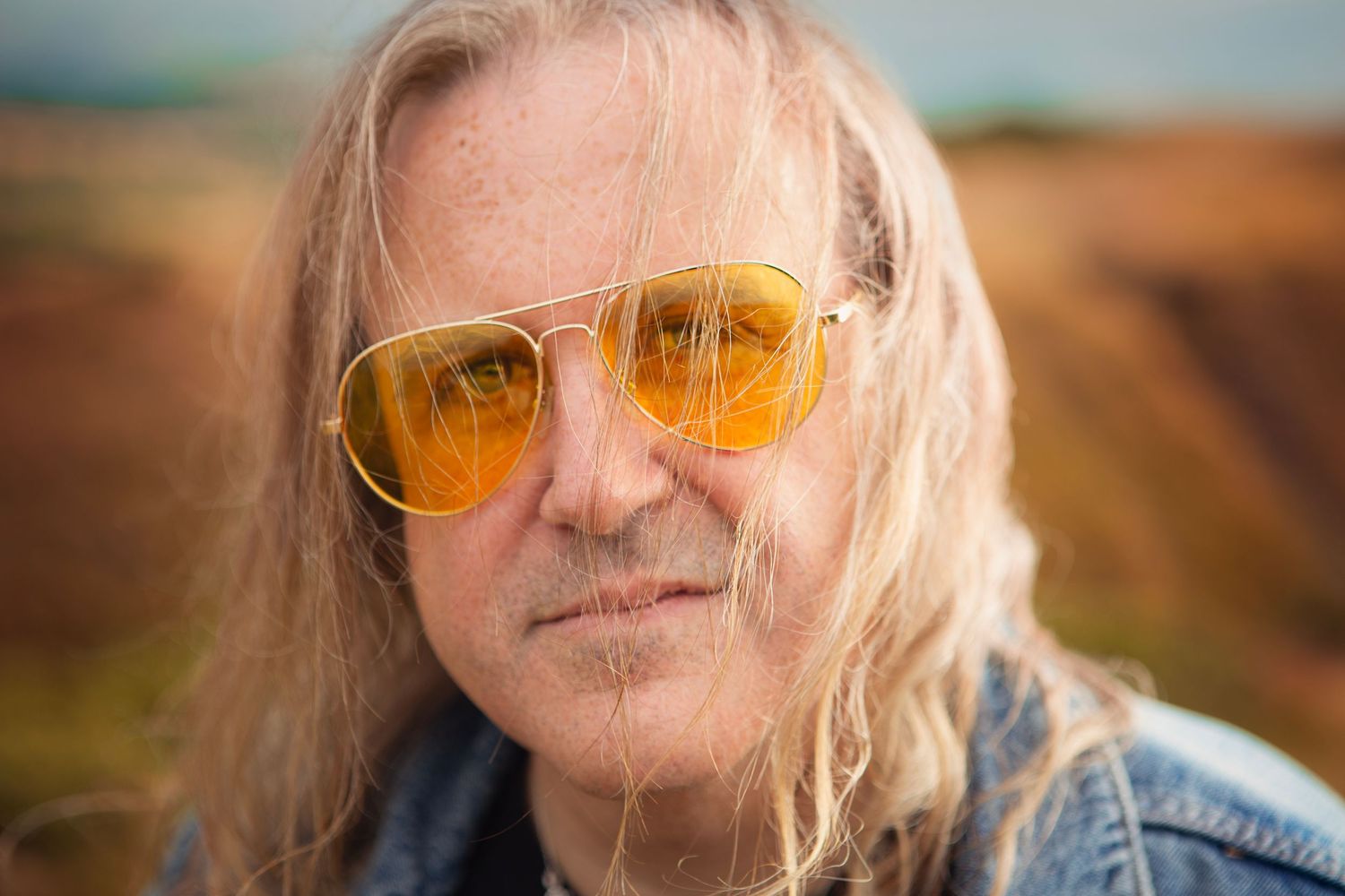 A long-haired older man wearing yellow tinted aviator sunglasses poses outdoors with a blurred landscape in the West Midlands