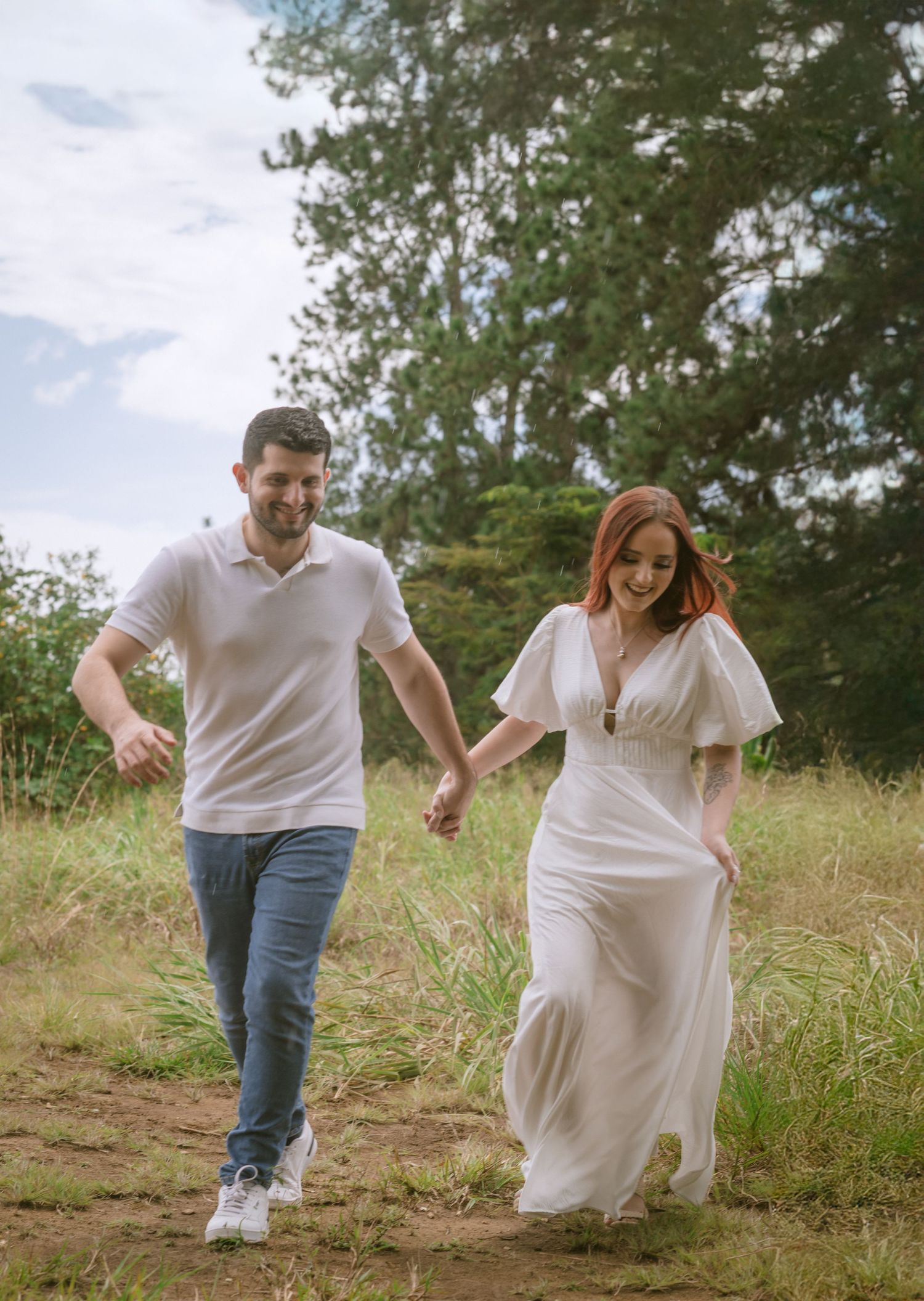 A couple runs hand in hand through a grassy hillside path wearing coordinating white outfits.