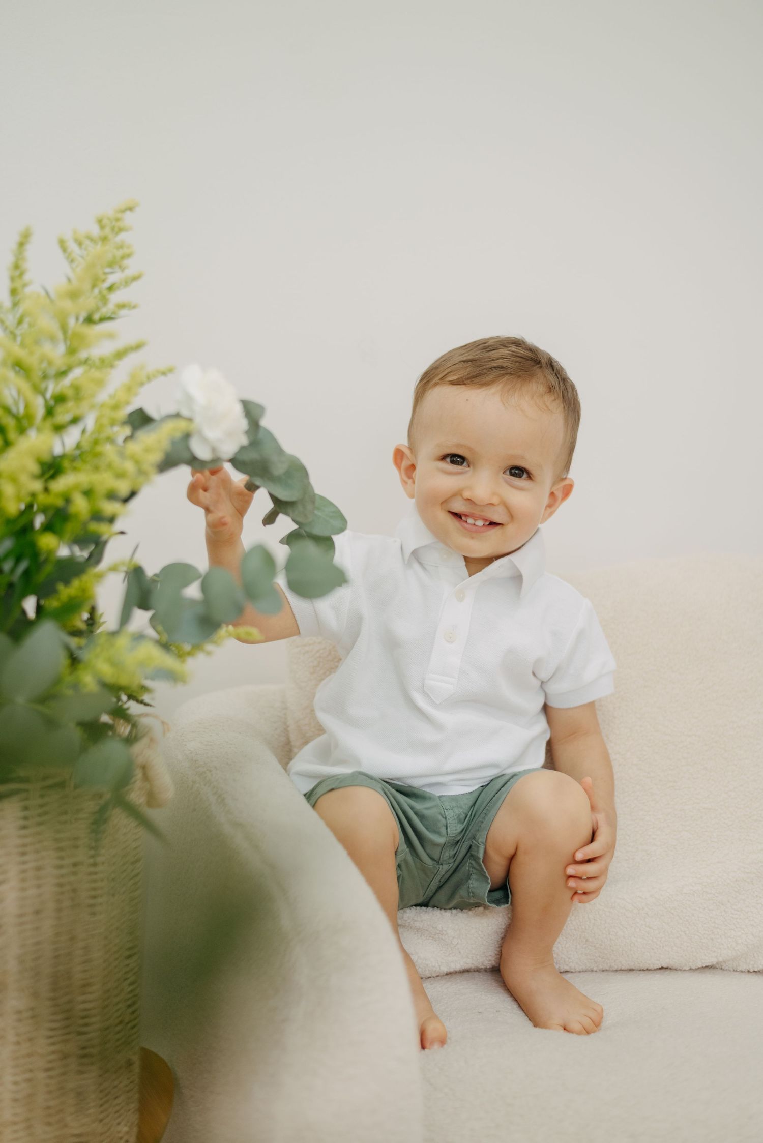 Charming portrait of toddler in white shirt sitting on cream couch next to greenery.