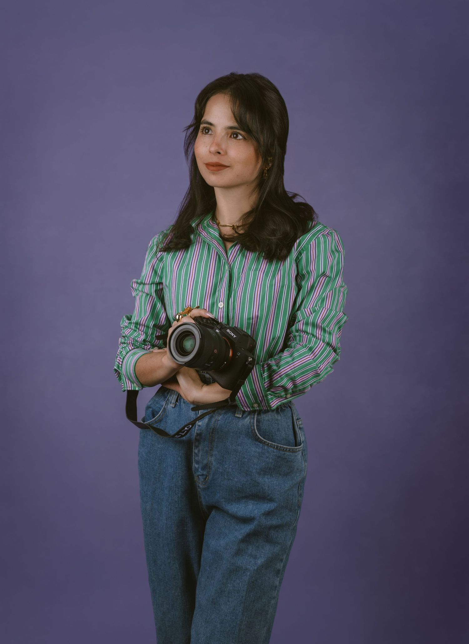 Photographer in striped shirt and jeans holds vintage camera against purple background.