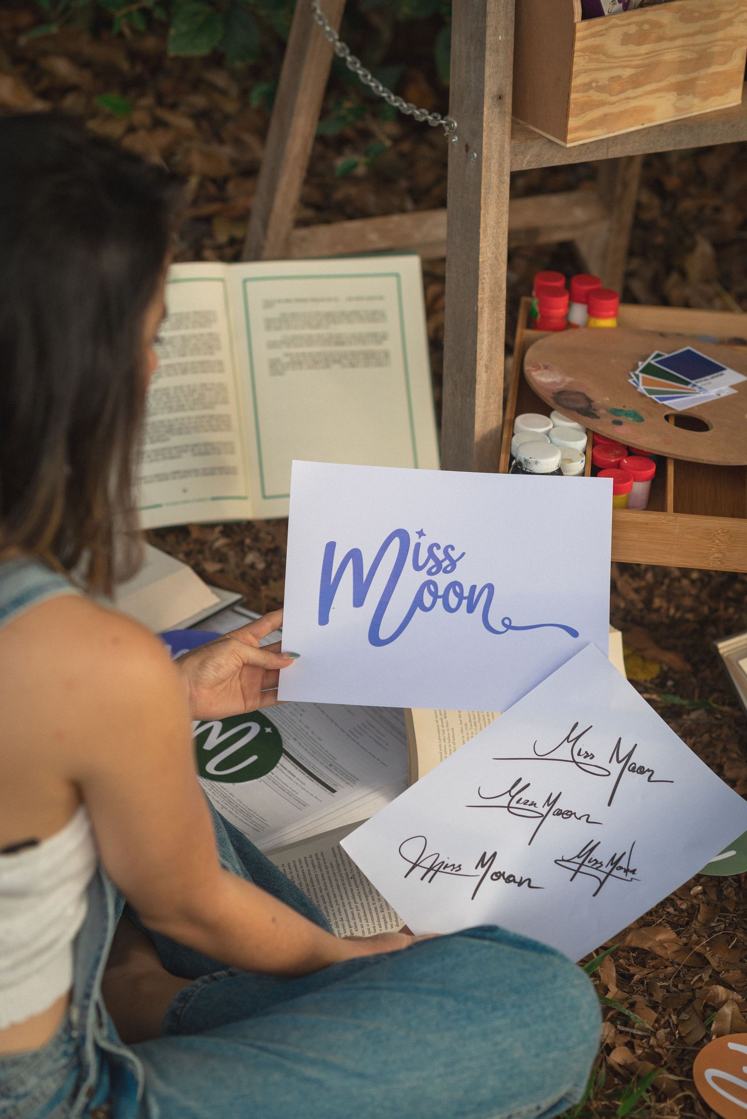 A person sitting on the ground practices calligraphy writing the word 'Moon' in blue ink with other papers nearby.