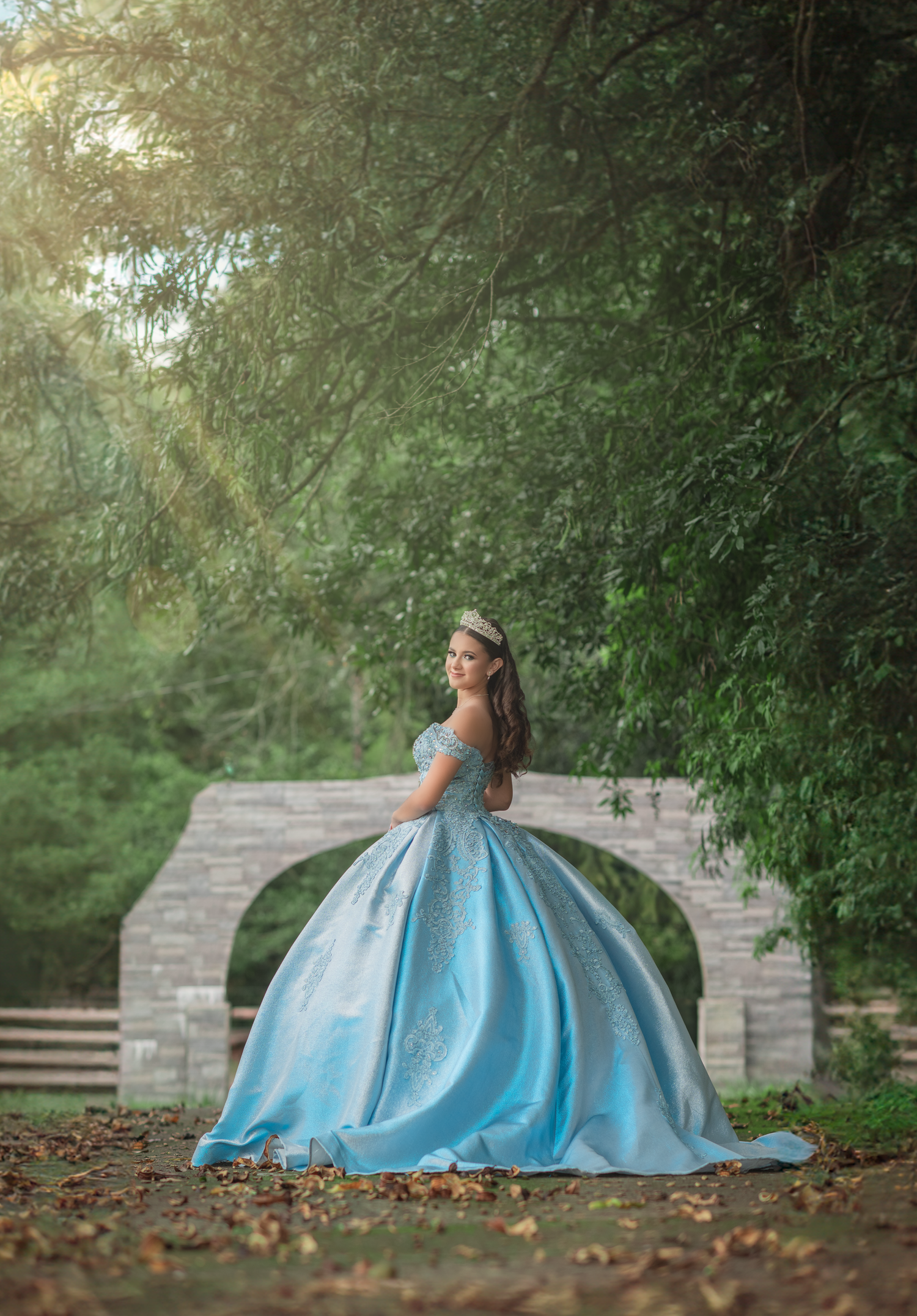 A flowing blue ball gown photographed against a stone bridge in a lush garden setting.
