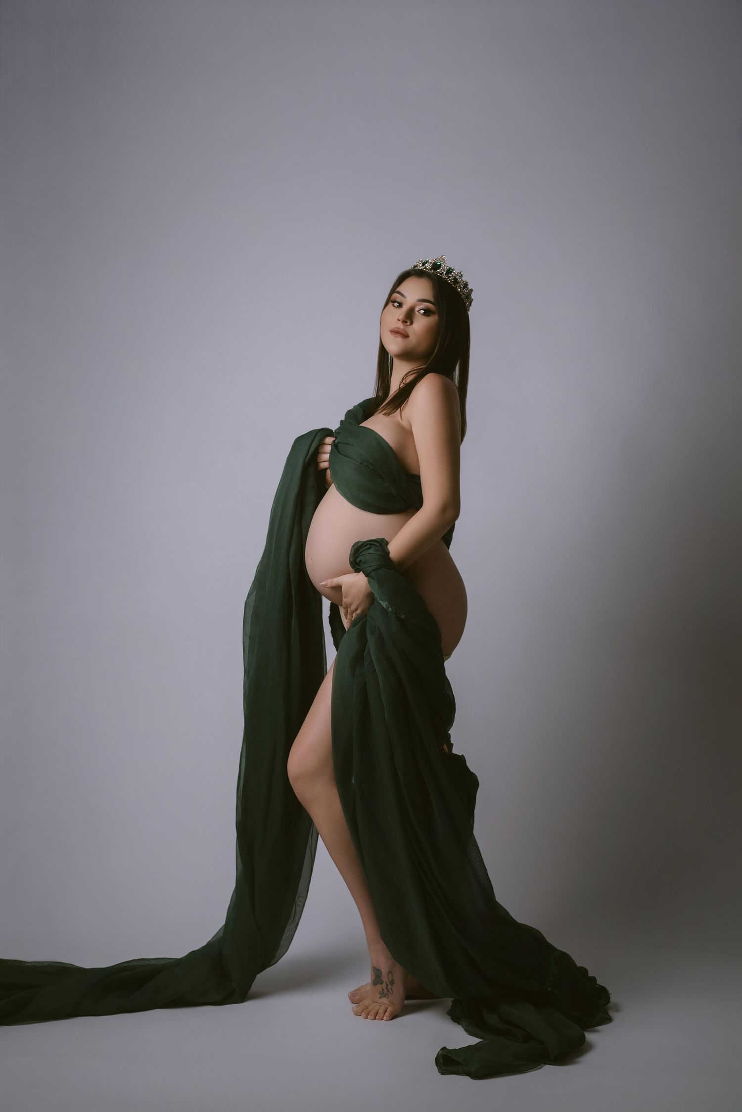 Dramatic maternity portrait with flowing green fabric and crown against neutral backdrop.