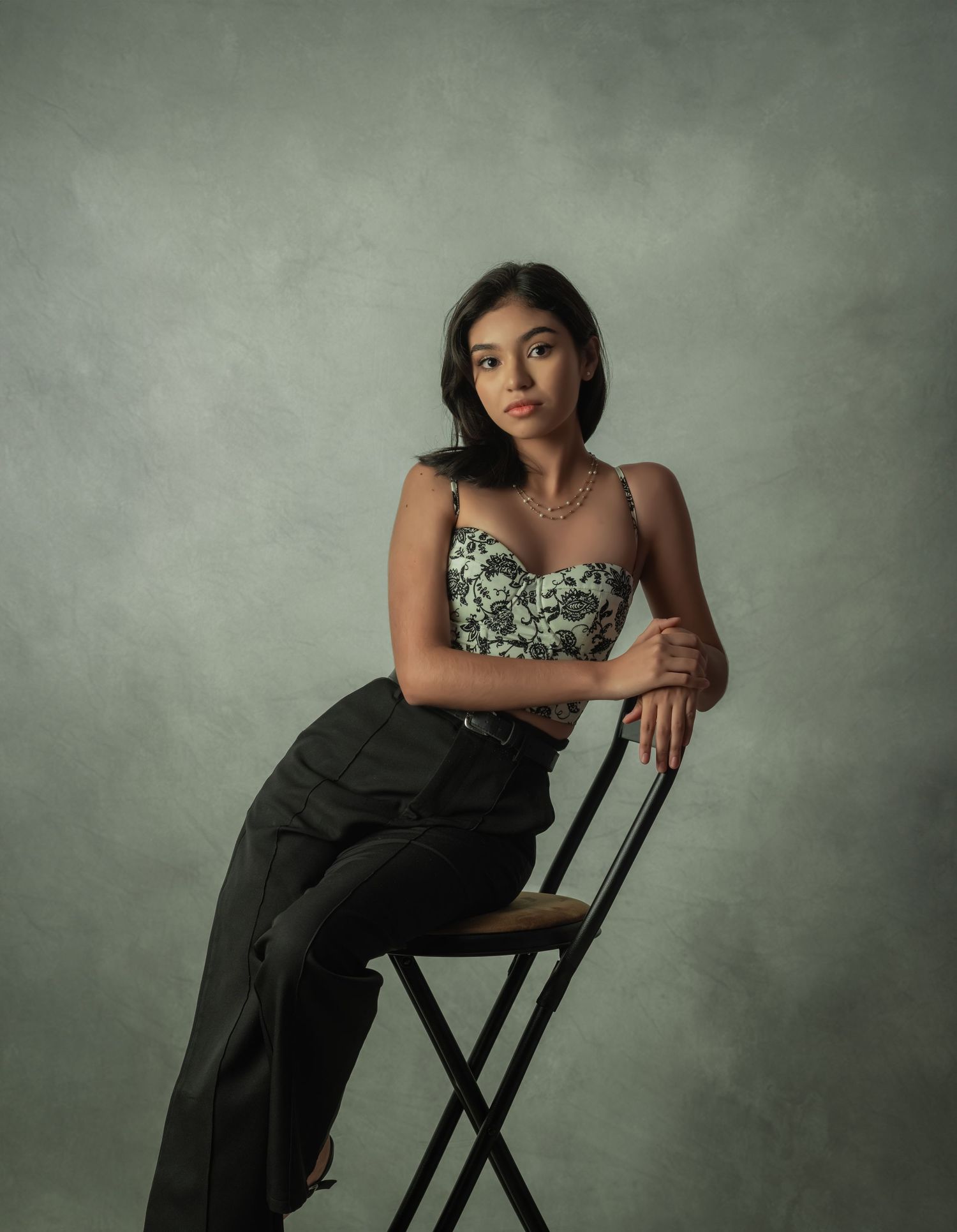 Studio portrait of a woman in floral top and dark pants posed on high chair.