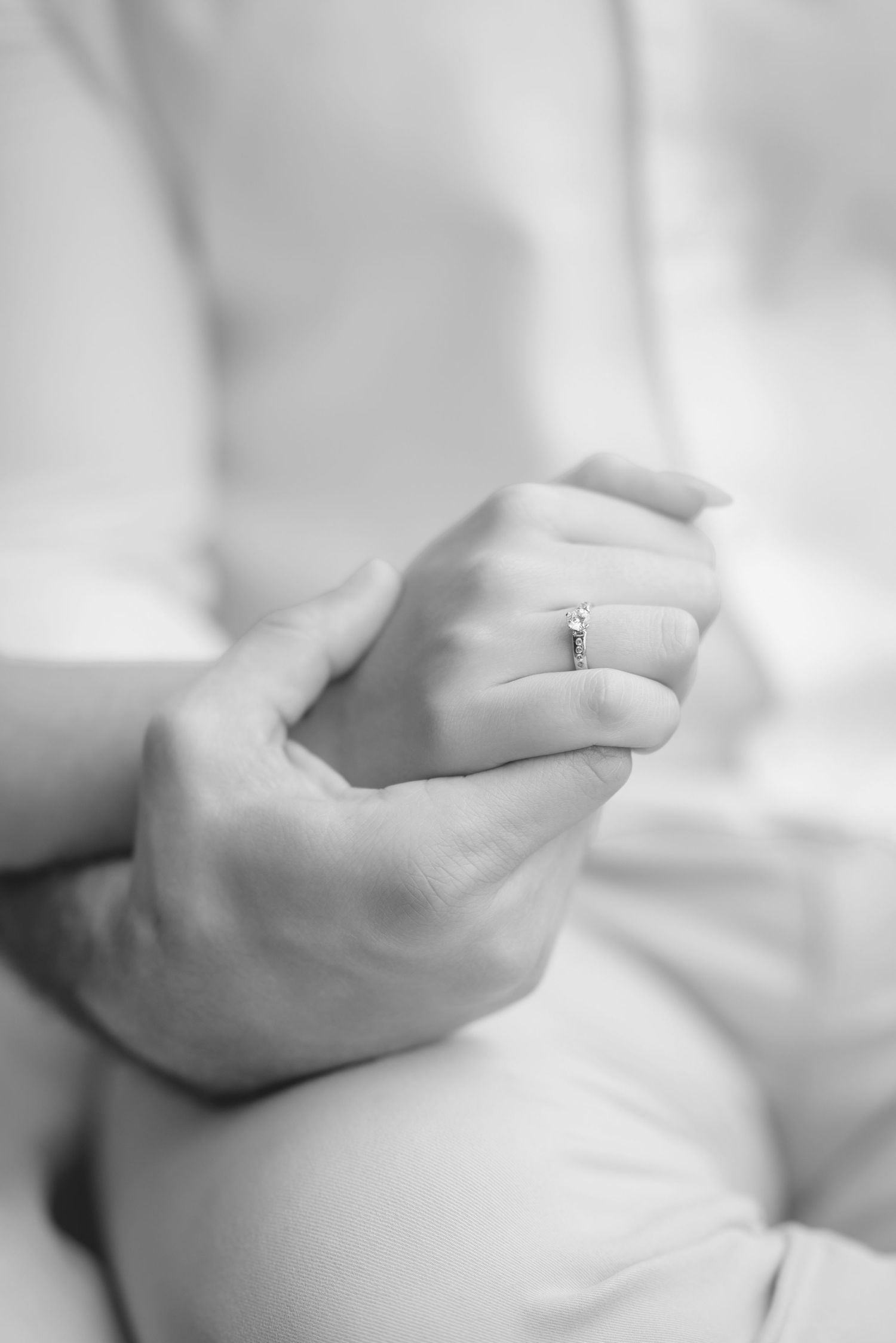 A black and white close-up of hands gently clasped together showing an engagement ring.