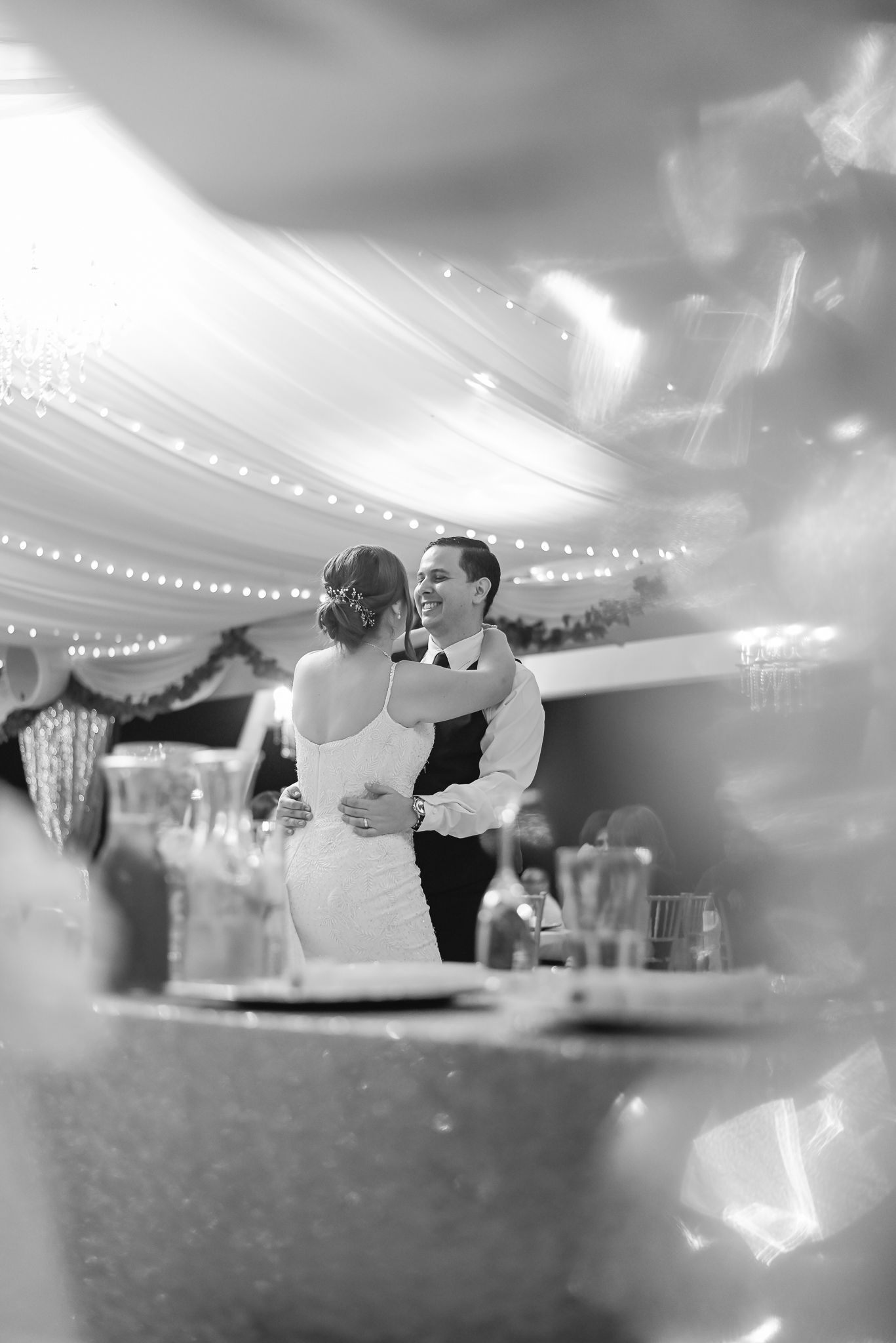 A romantic black and white photo of a couple's first dance at their wedding reception under string lights.
