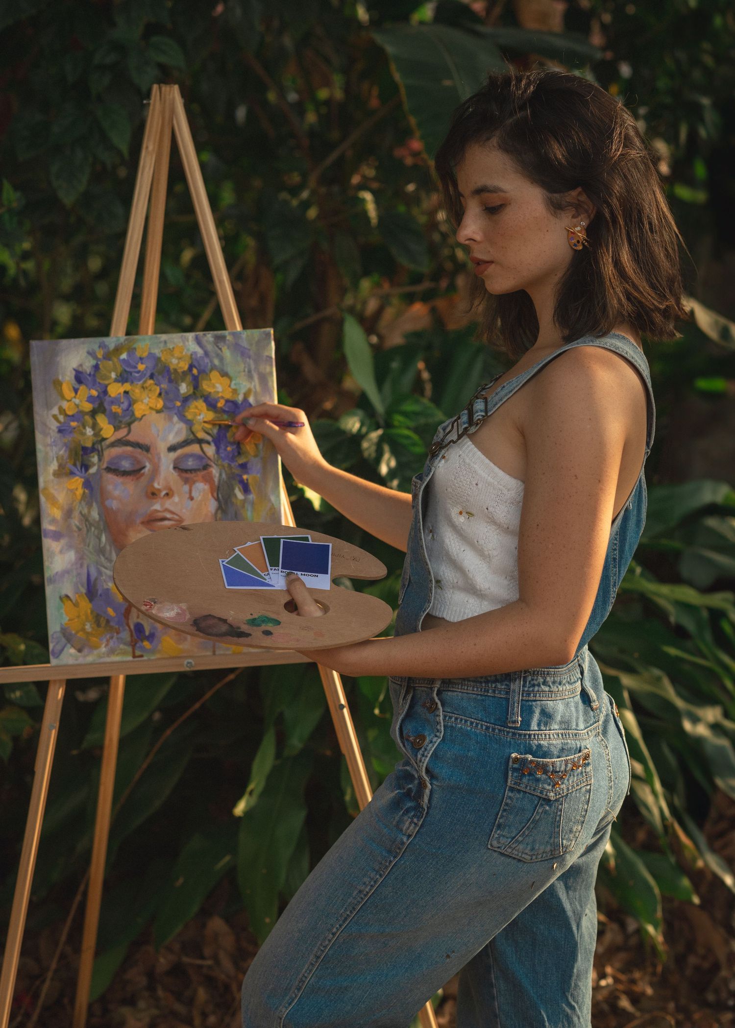 Artist painting a portrait of a mystical figure with purple flowers and holding cards while standing at an easel outdoors.