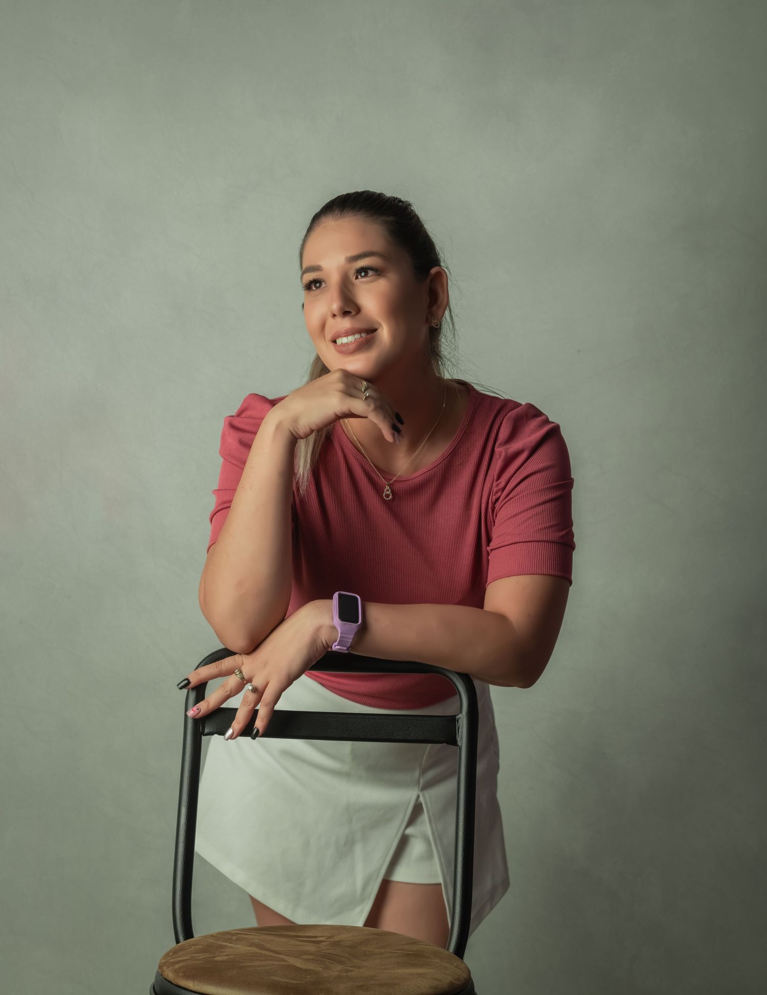 A woman in a pink top poses thoughtfully against a gray backdrop while leaning on a chair.