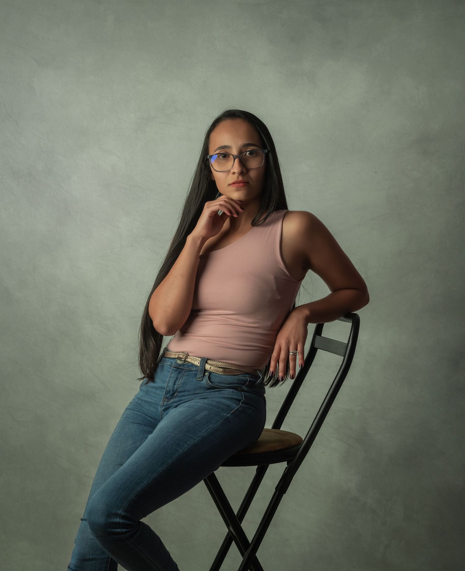 Studio portrait of a woman in pink top and jeans posing on chair.