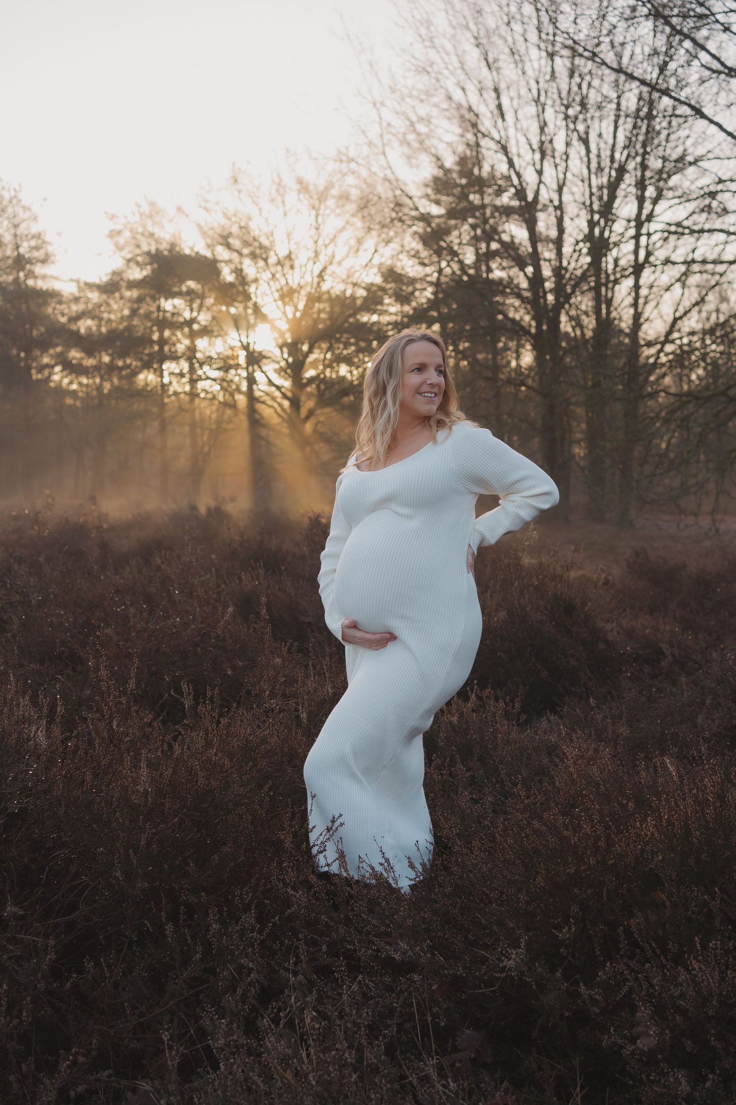 Zwangere vrouw in witte jurk tijdens een zwangerschapsshoot op de heide bij zonsopgang