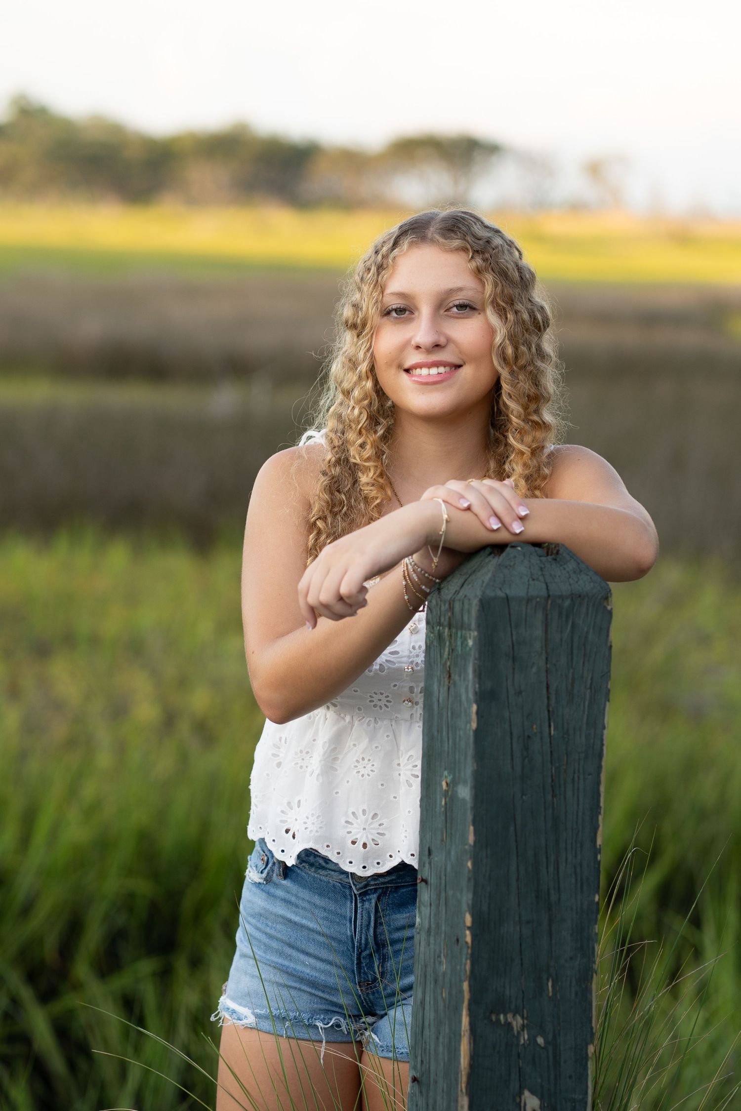 Smiling teenage girl with curly blonde hair leans on a wooden post in a field wearing a white top and denim shorts.