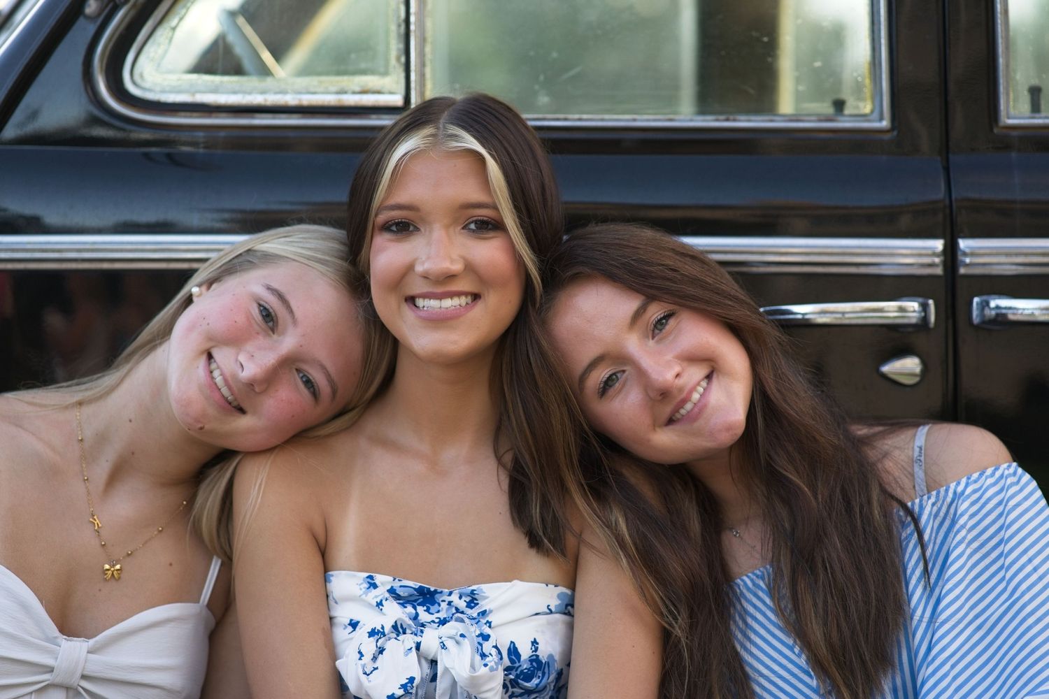 Three teenage girls smiling together in front of a vintage black car in summer outfits.