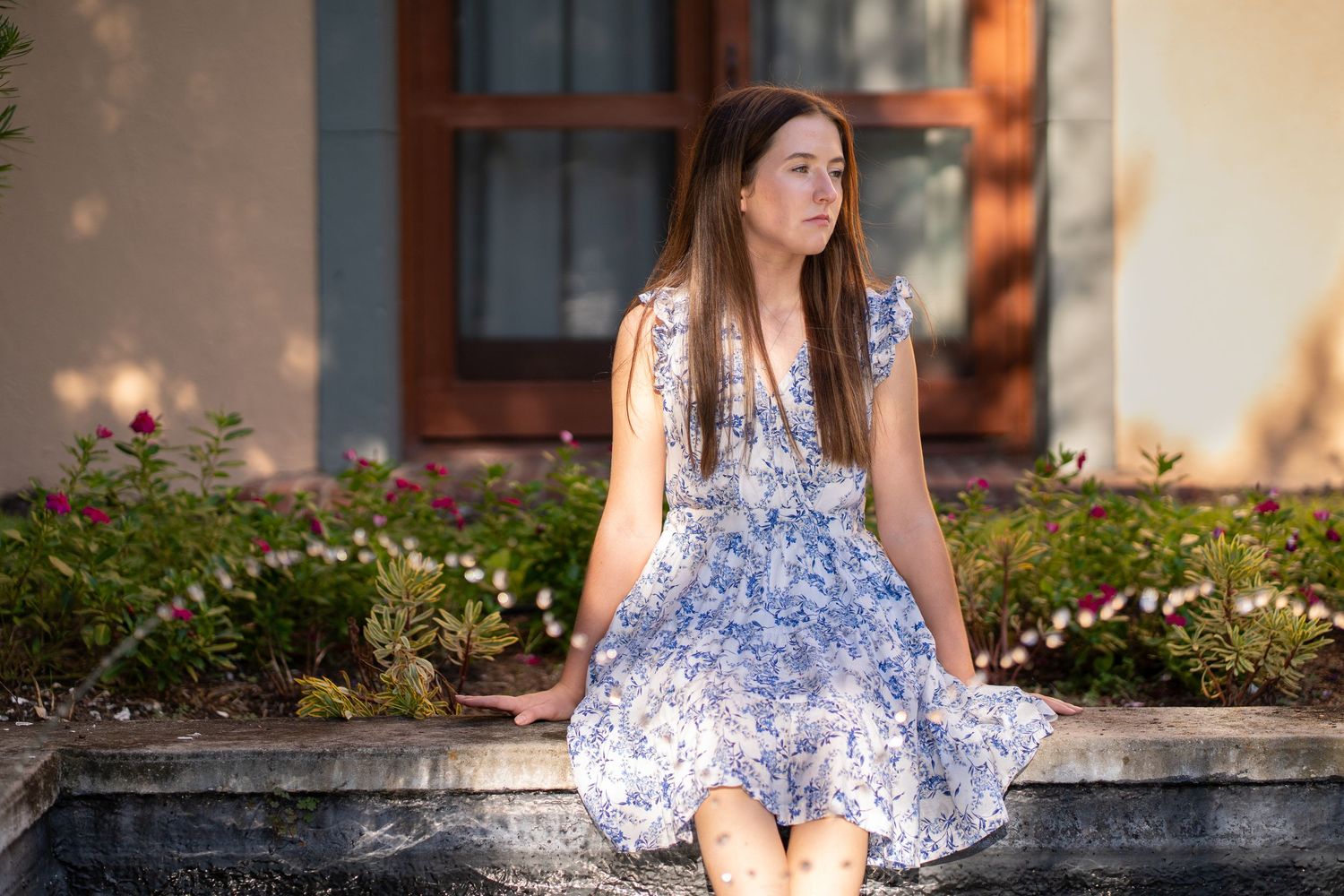 Young woman in blue floral dress sitting on stone fountain edge surrounded by garden flowers.