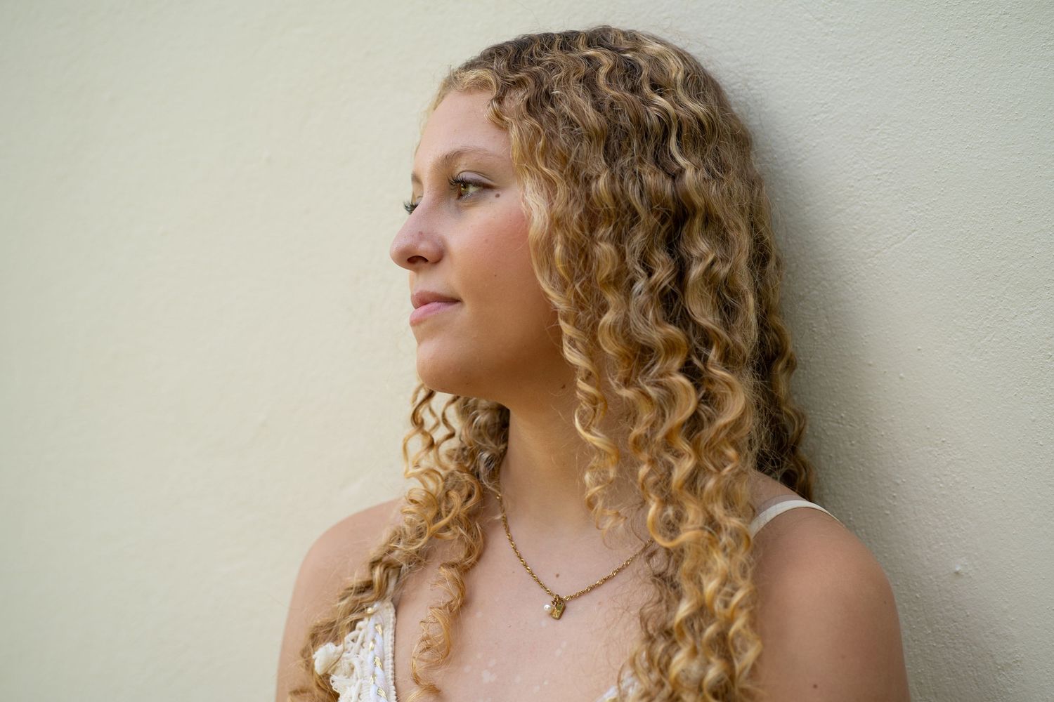 Young woman with long curly blonde hair wearing a delicate necklace, posing against a light green wall.
