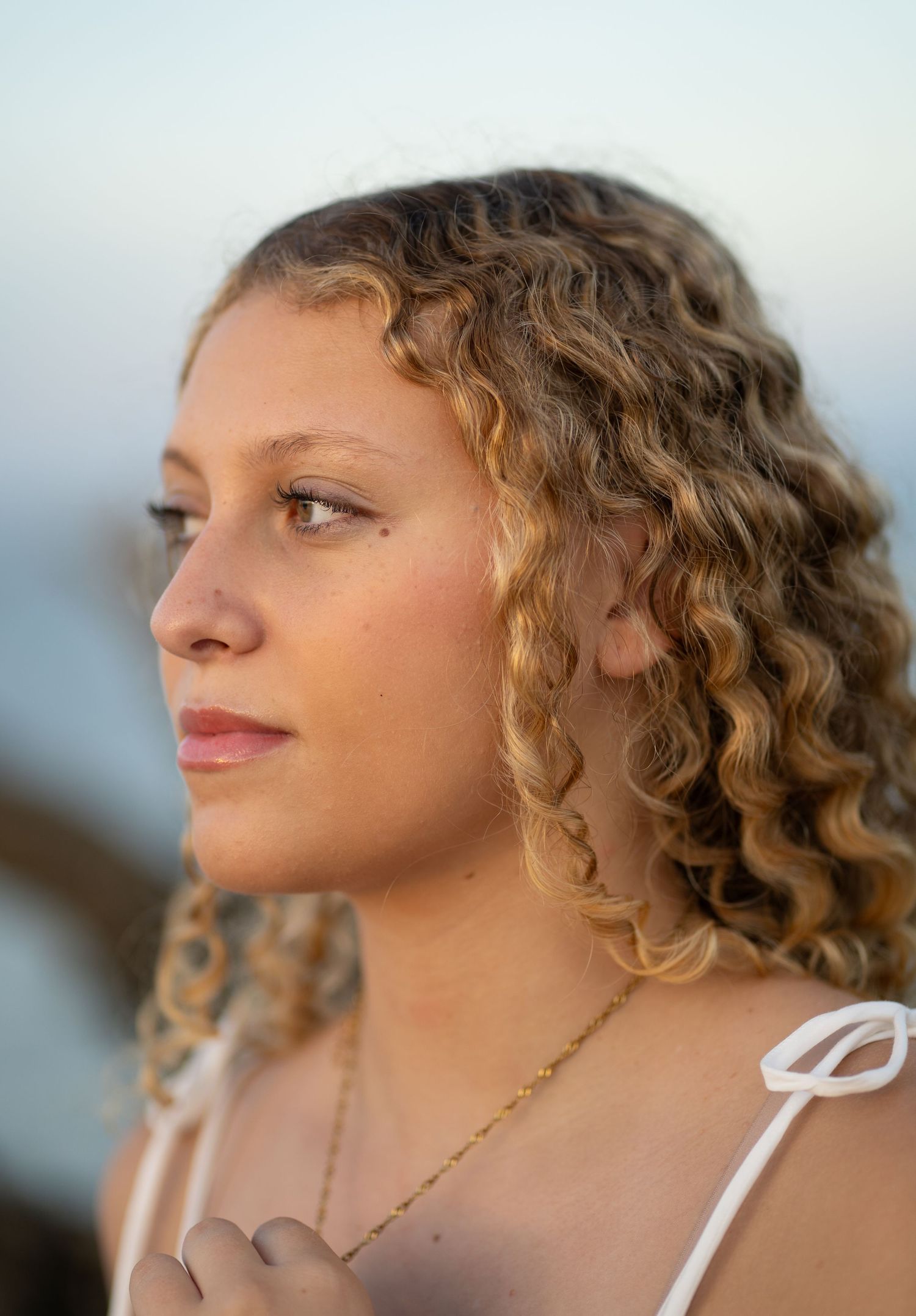 Young woman with curly blonde hair wearing a white top and gold necklace, gazing thoughtfully in natural light.