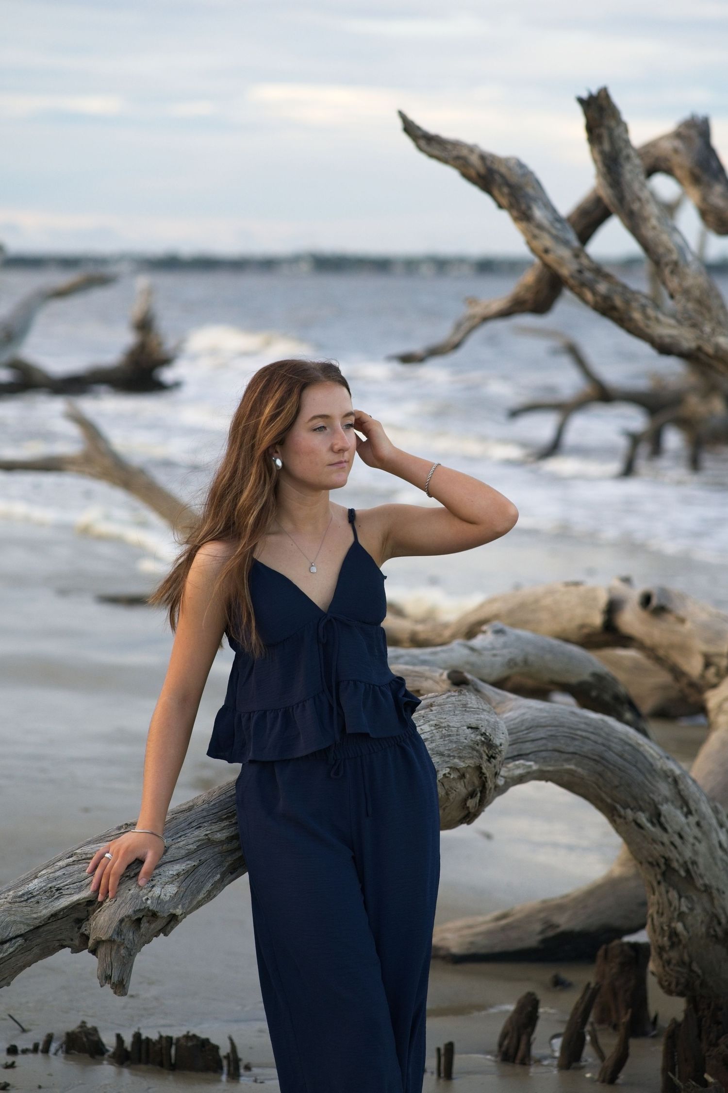 Senior portrait of a young woman in navy outfit leaning on driftwood at beach, gazing away with ocean waves behind her.