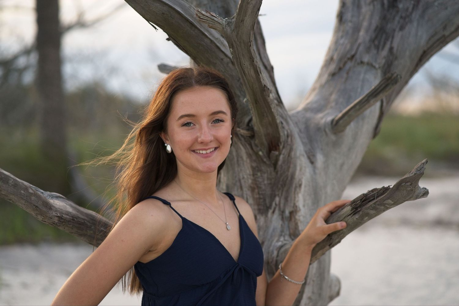 Senior portrait of a smiling teenage girl in navy dress posing beside a weathered driftwood tree on the beach.