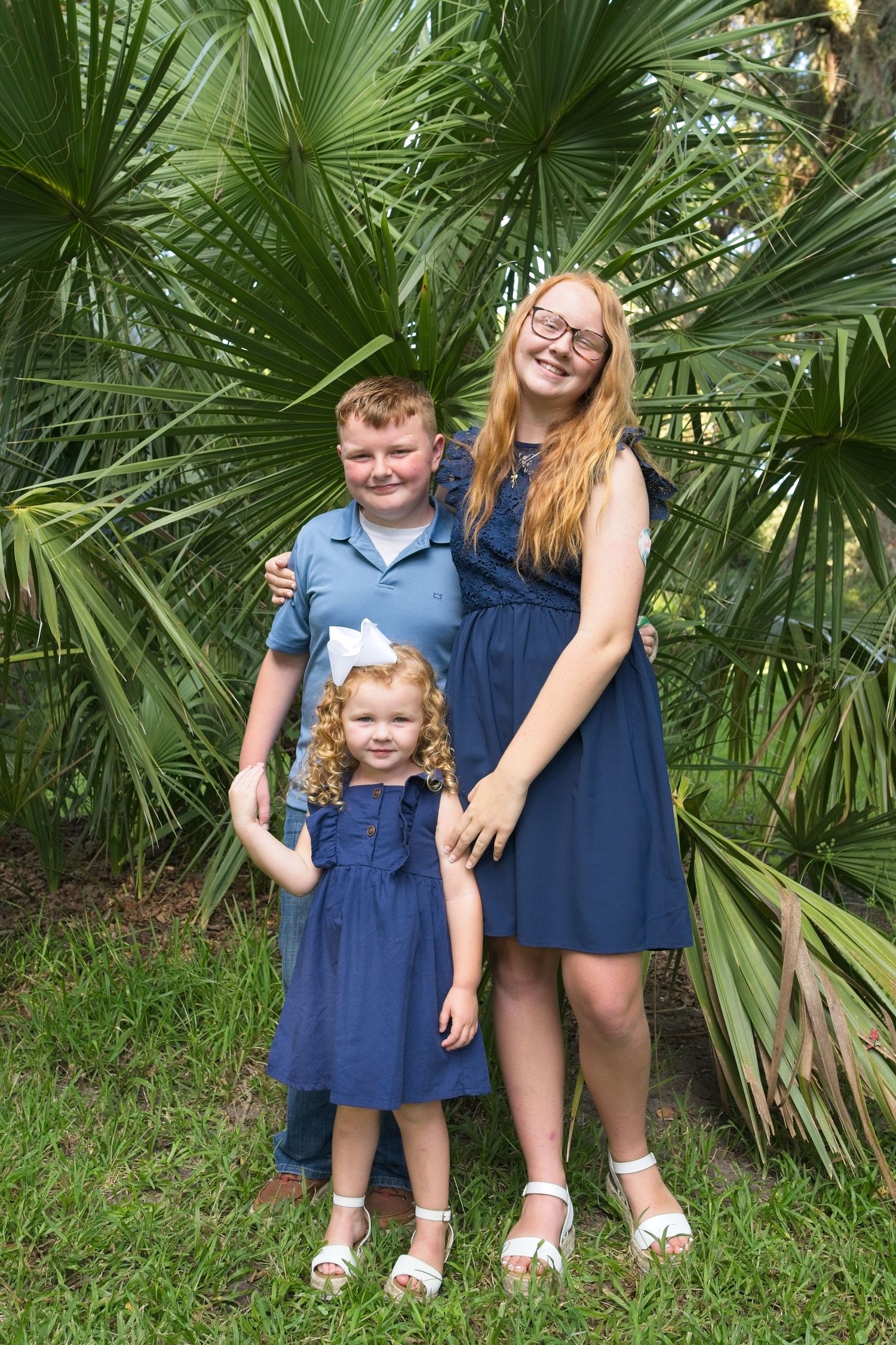 Three siblings in coordinating blue outfits smiling together in front of lush tropical palm trees.