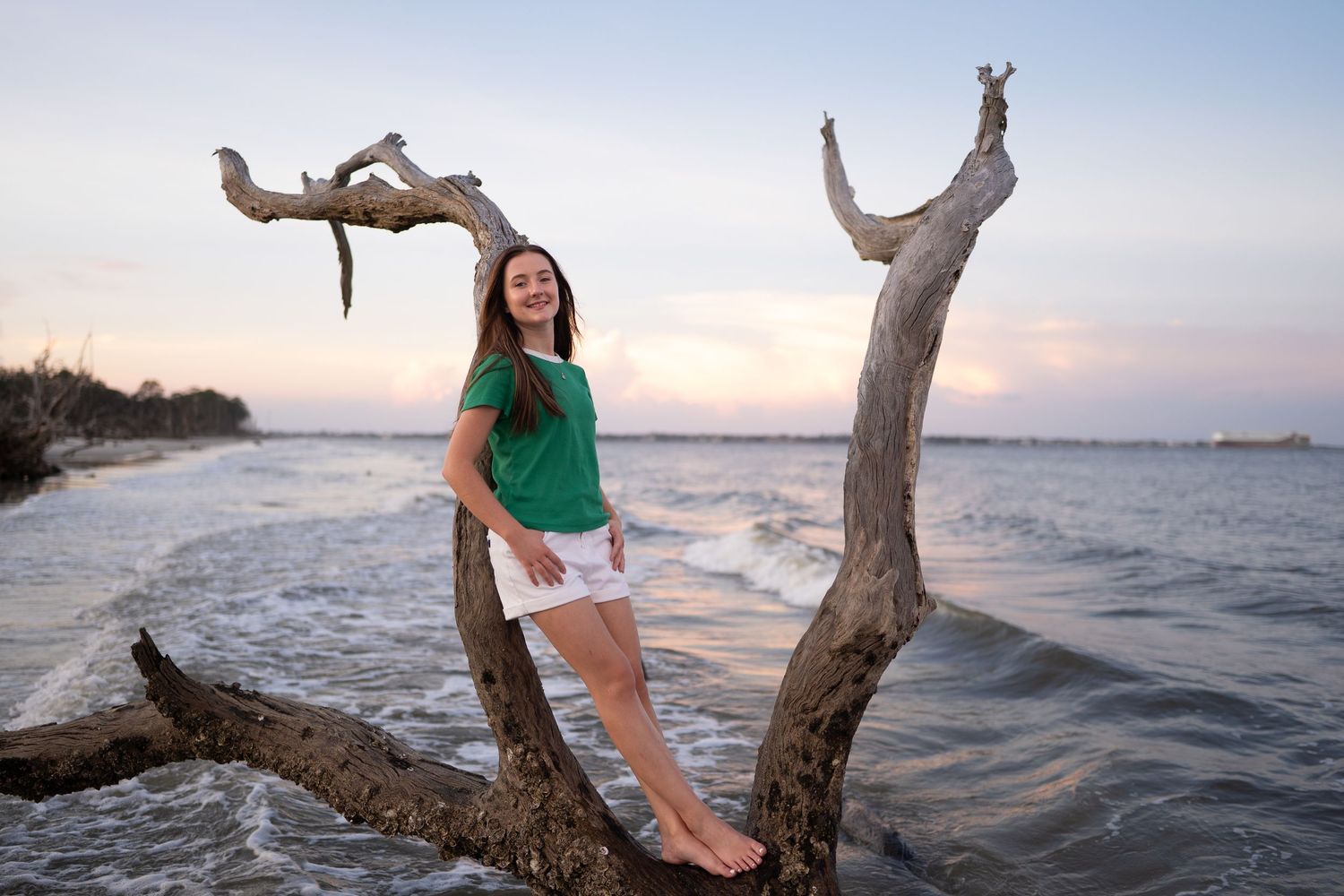 Teen girl in green top and white shorts poses on driftwood at a beach during a colorful sunset.