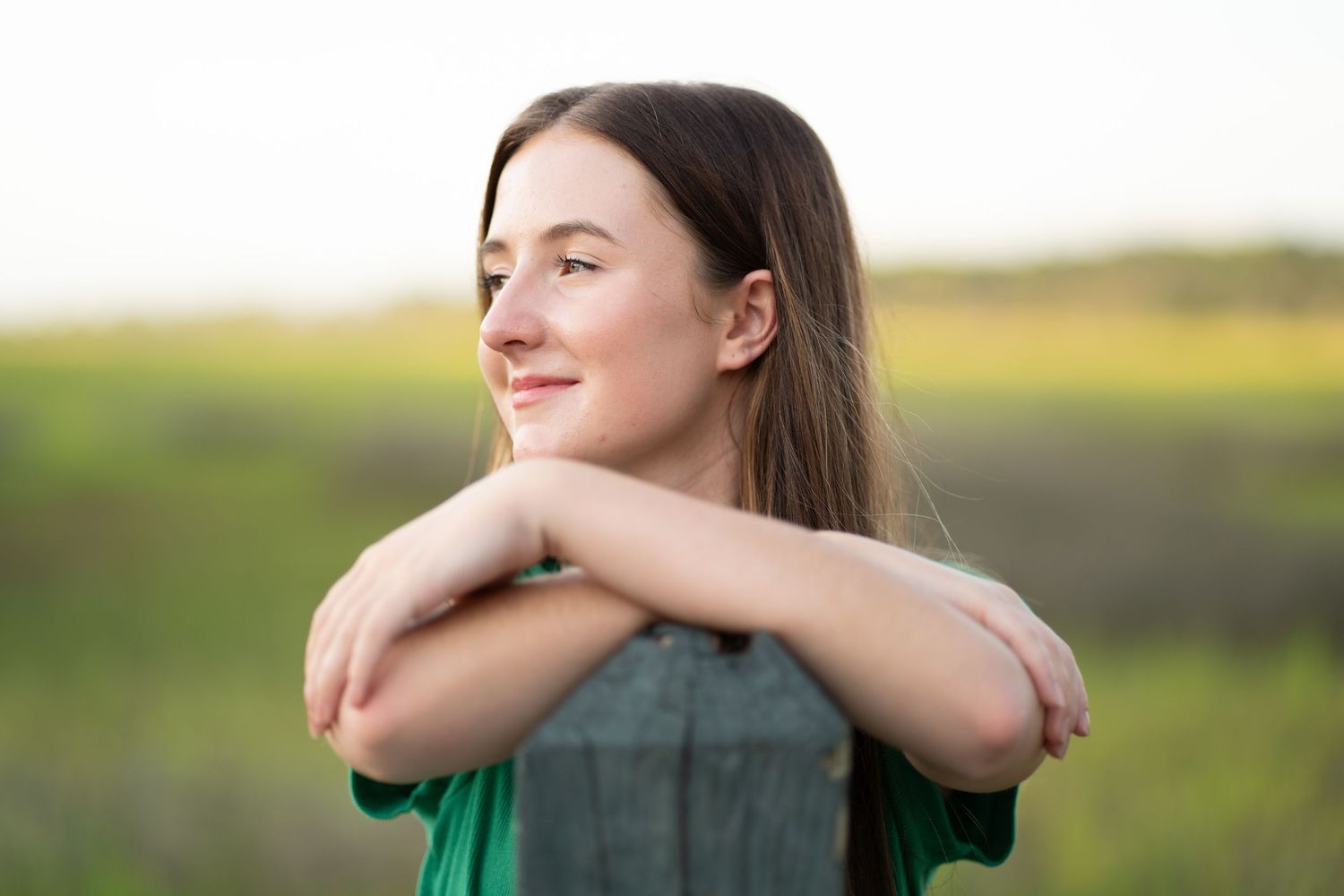 Young woman in green shirt smiling outdoors, arms crossed on fence post, with green fields in background.