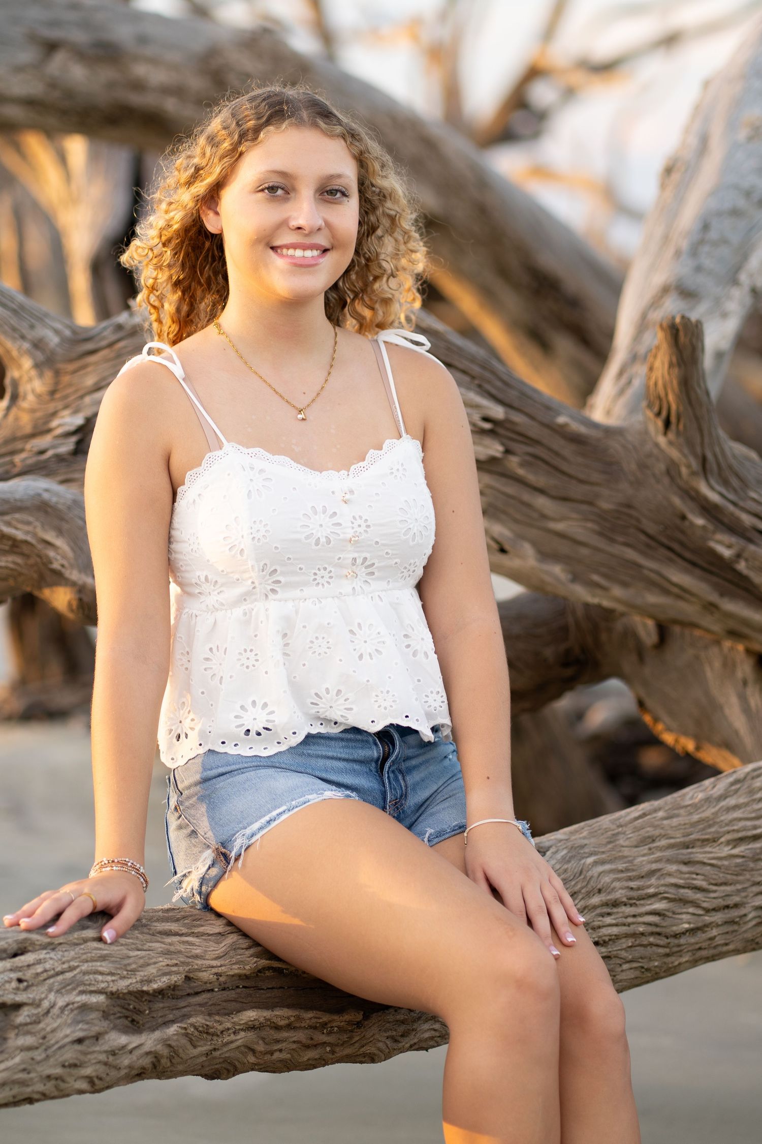 Young woman with curly hair sits on driftwood at the beach, wearing a white eyelet top and denim shorts, smiling.