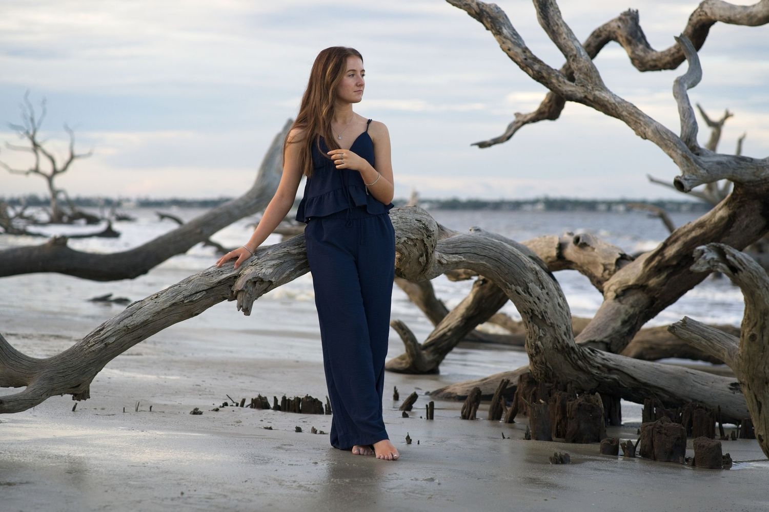 Senior portrait of a young woman in navy jumpsuit posing gracefully against large driftwood on a scenic beach at dusk.
