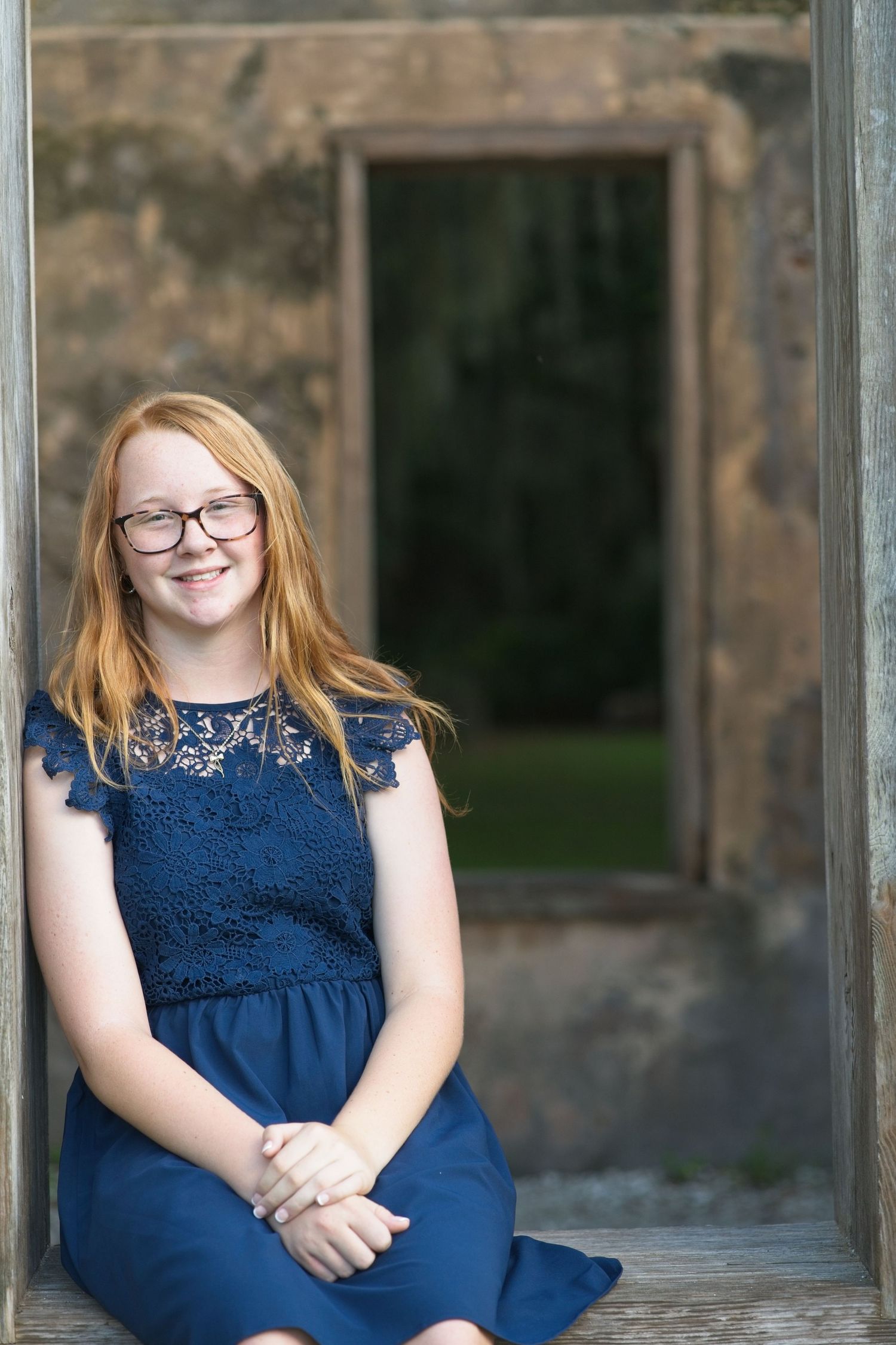 Smiling red-haired teenage girl in navy lace dress sitting in a rustic stone doorway.