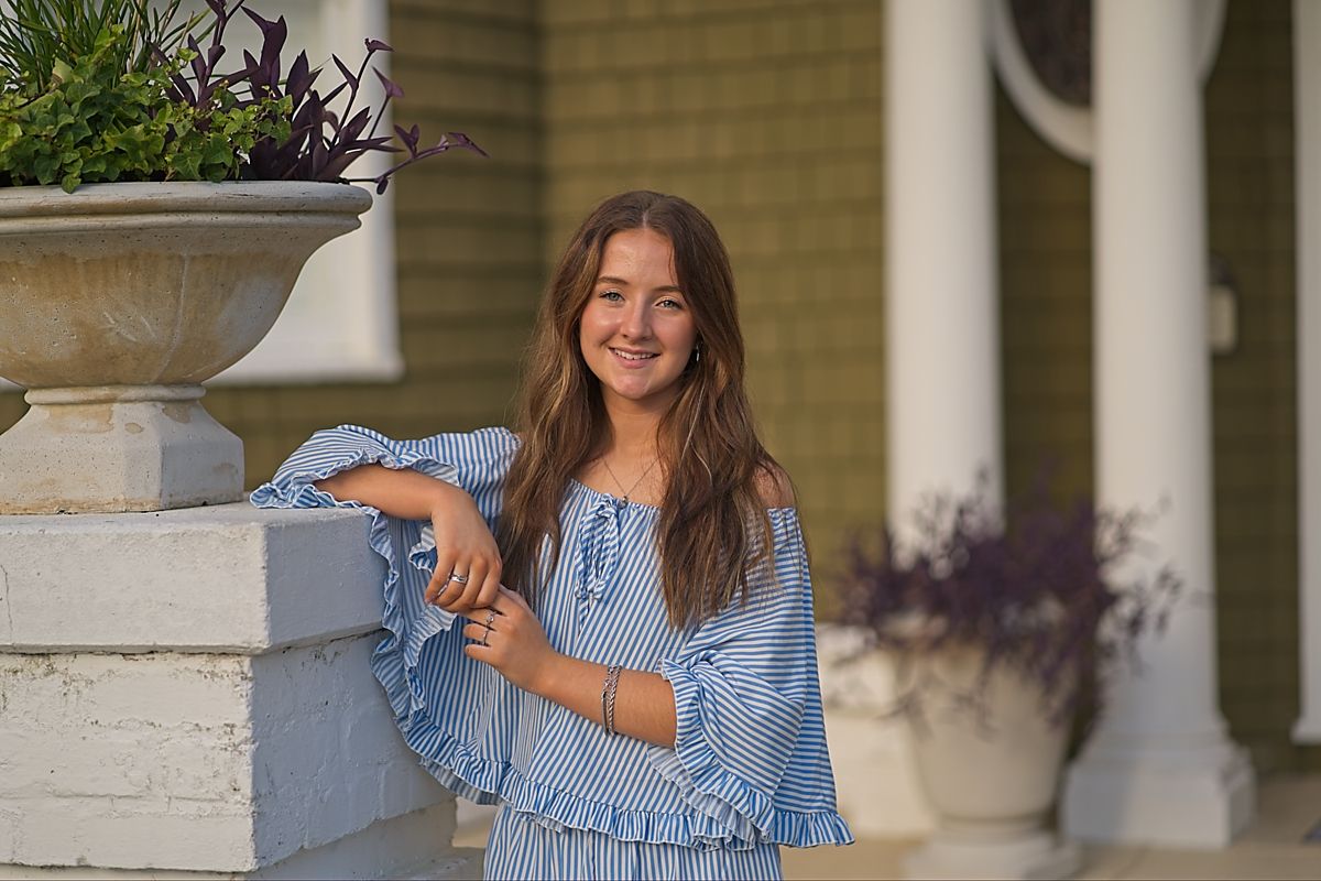 Senior portrait of a young woman in blue striped off-shoulder dress posing by white pillar with decorative planter outdoors.