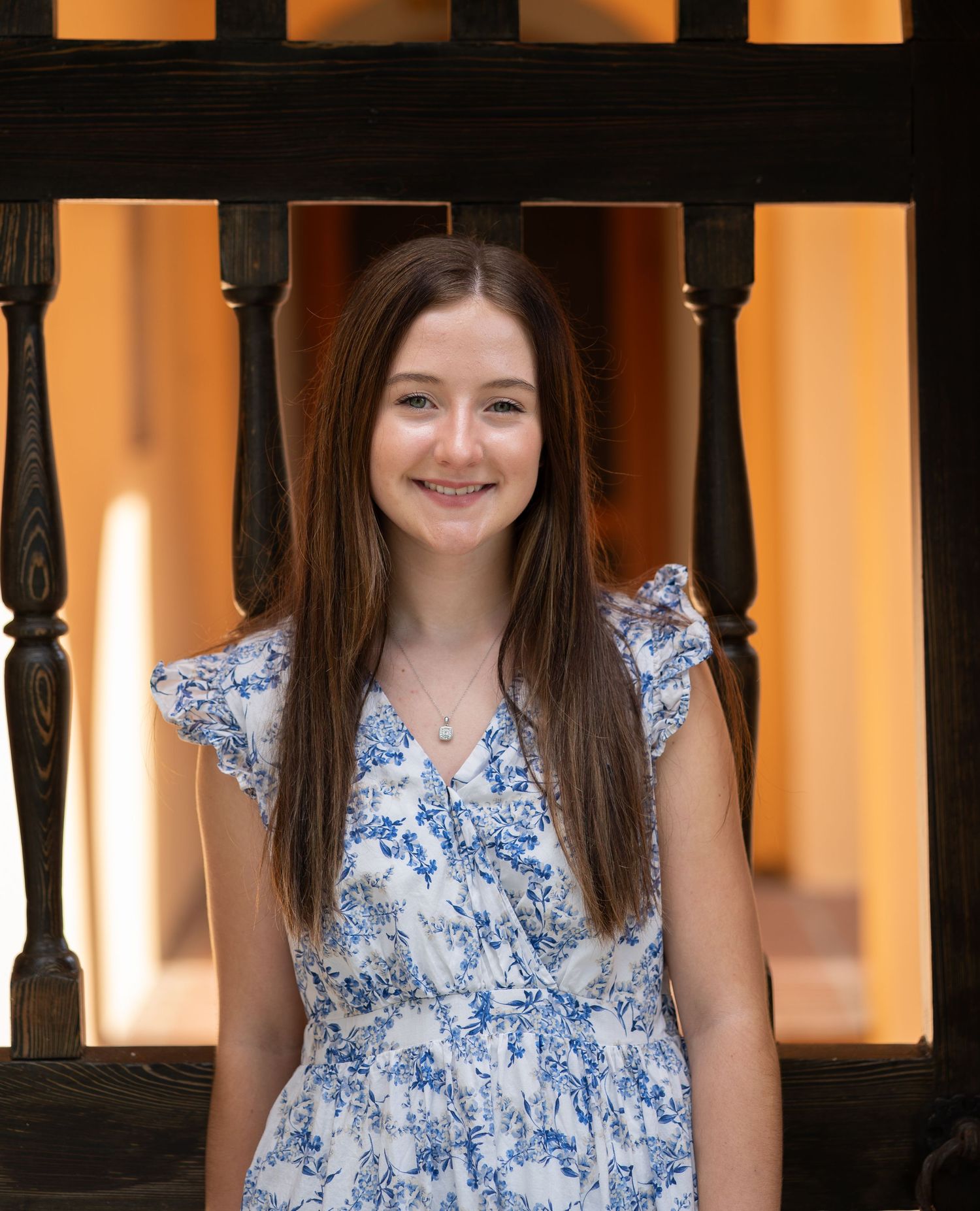 Smiling young girl with long brown hair wearing a white and blue floral dress, standing in front of dark wooden railings.