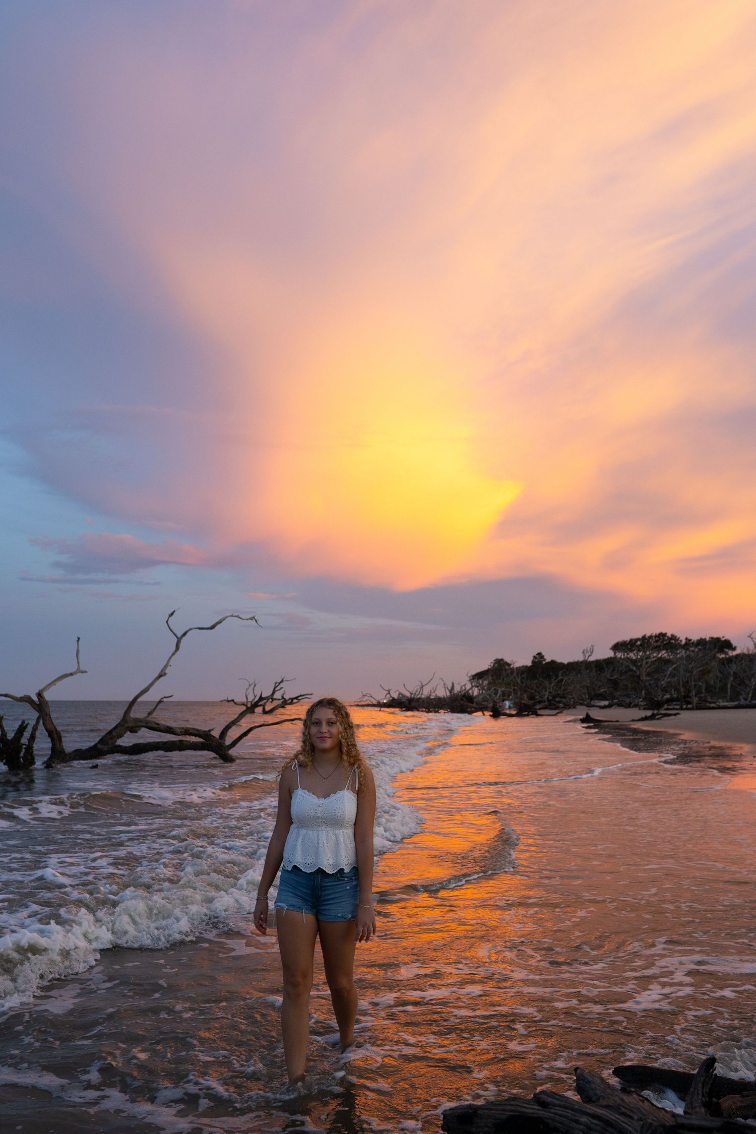 Young woman in white top and denim shorts standing on a beach at sunset with vibrant orange and pink sky.