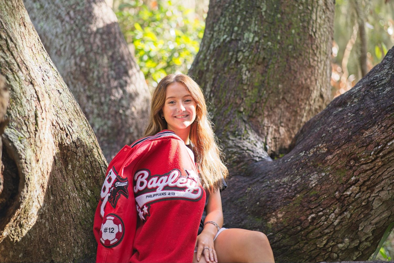 Senior portrait of a smiling teen girl wearing red Eagles baseball jersey posing between large trees outdoors.