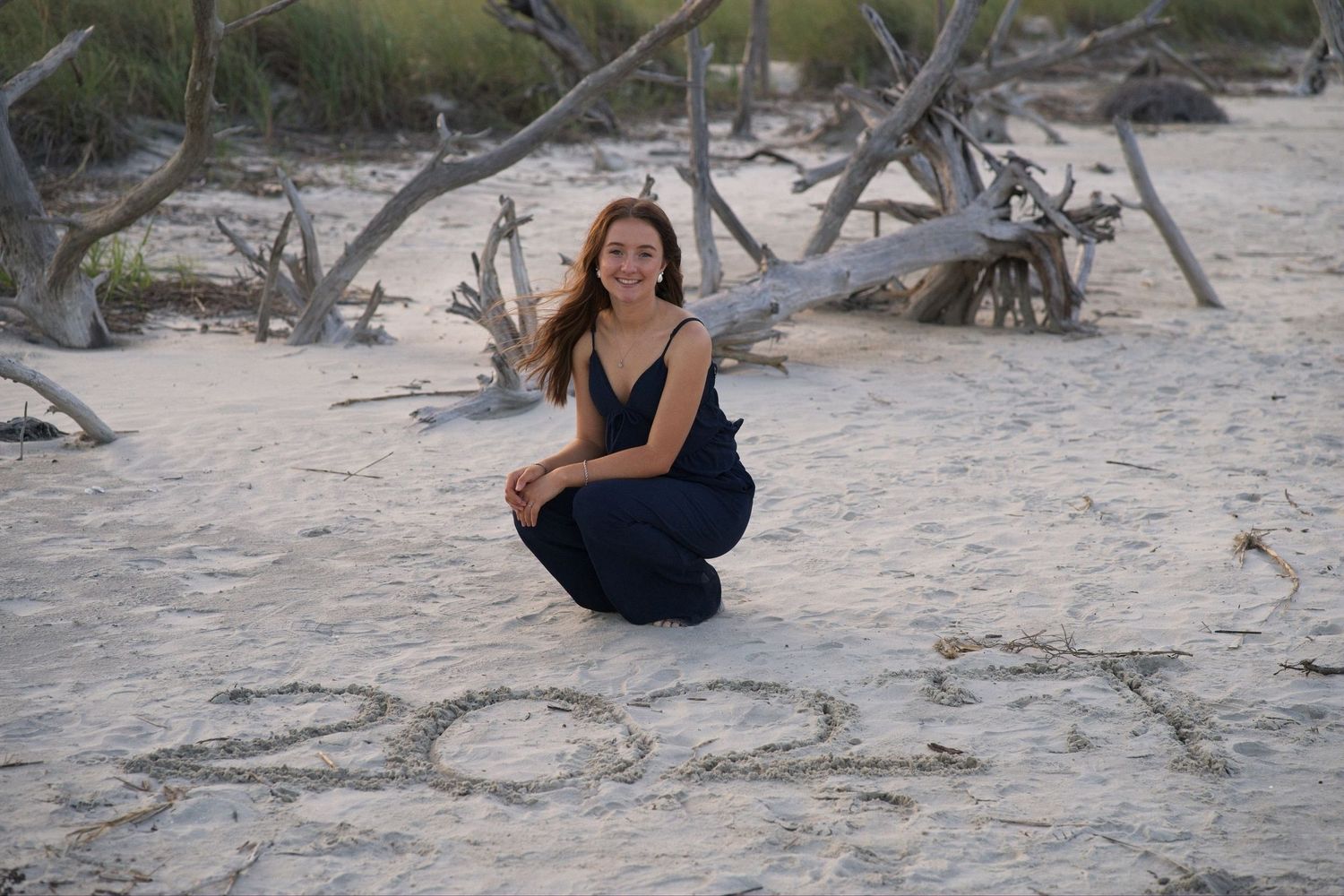 Senior portrait of a young woman kneeling on beach sand beside the year 2021 written in sand near driftwood.