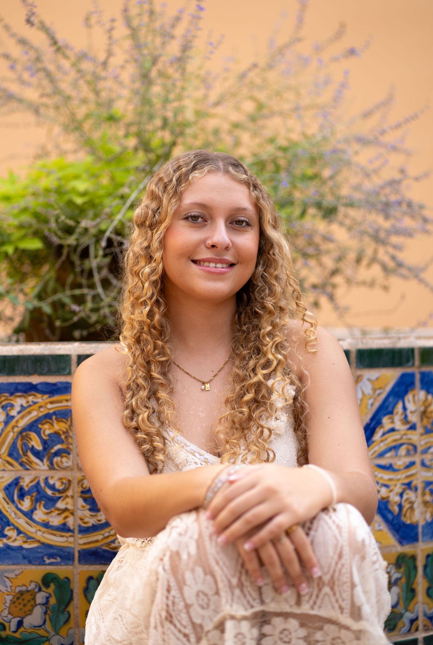 Young woman with curly blonde hair smiling, seated on decorative tiled bench with lavender plants behind her.