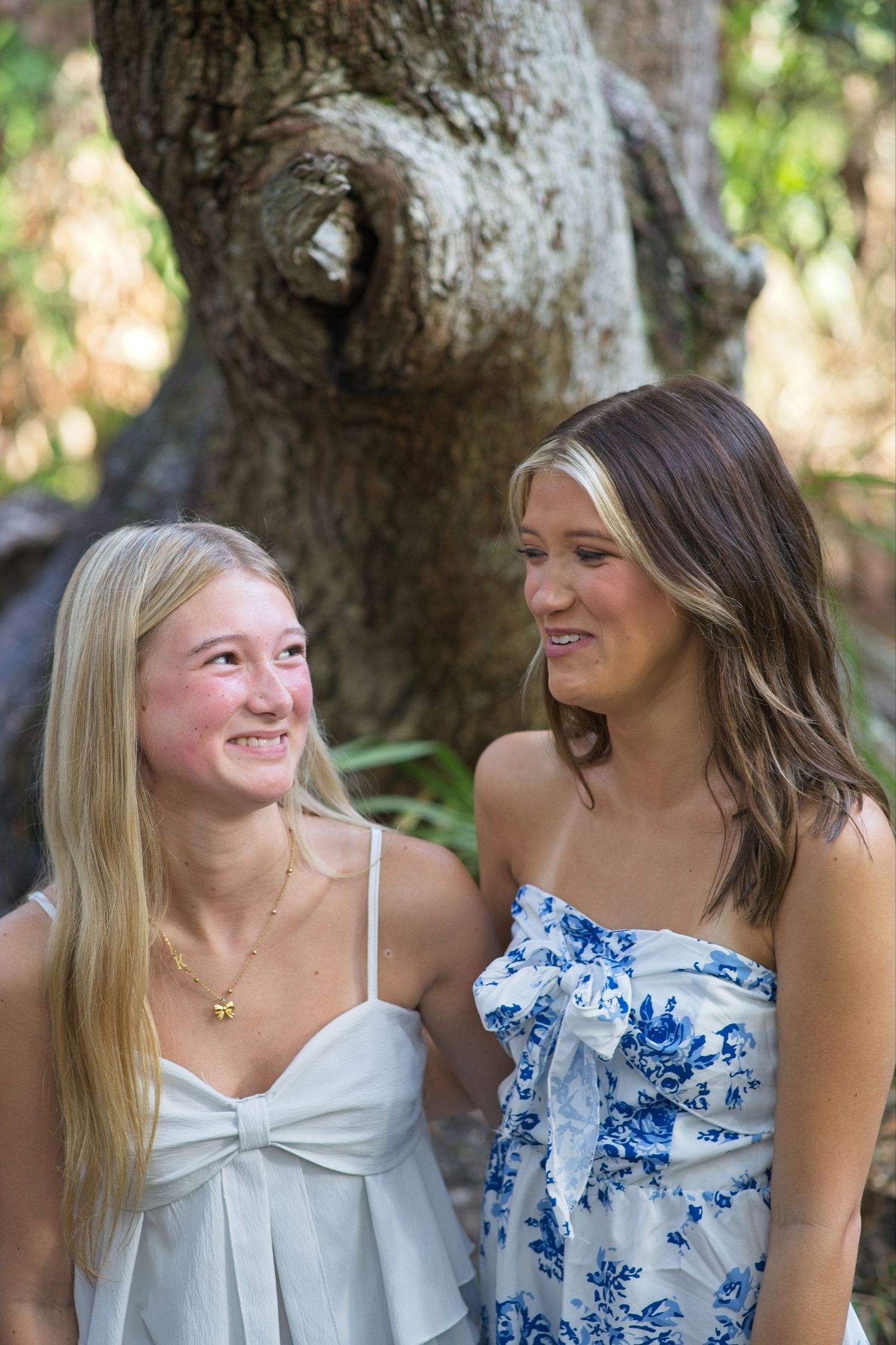 Two teenage girls laughing together in front of a large tree in a natural outdoor setting.