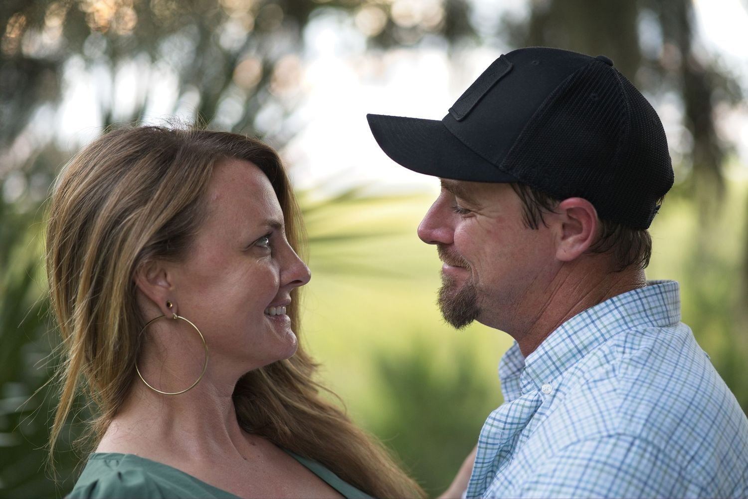 Couple smiling at each other outdoors with lush green trees in the background.