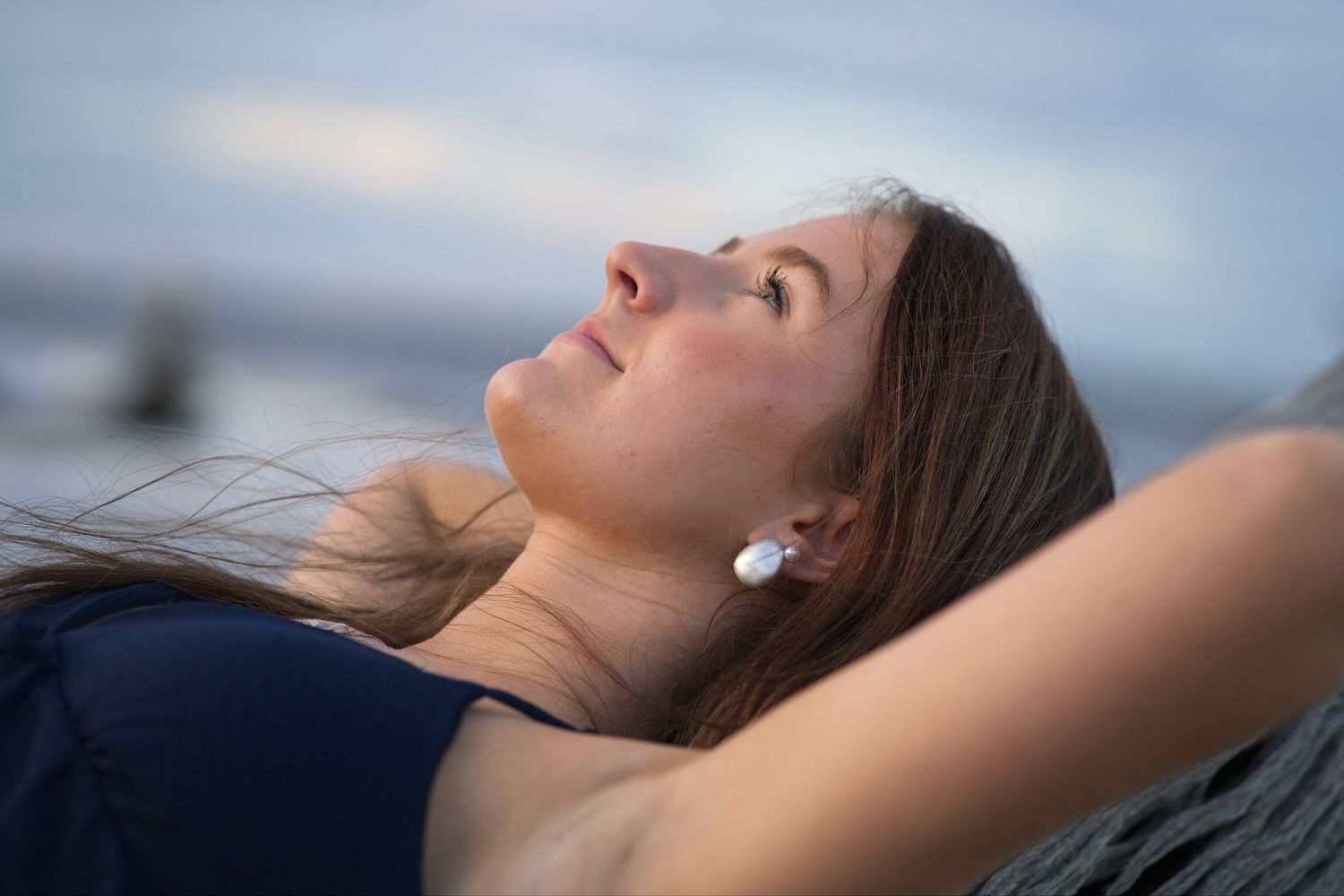 Senior portrait of a young woman with brown hair relaxing on driftwood wearing pearl earring gazing upward at sky.